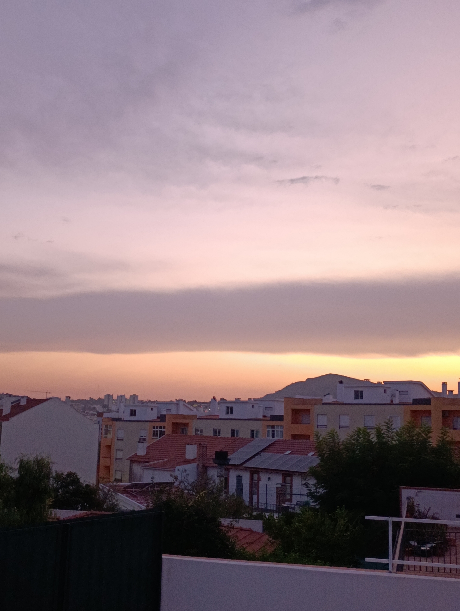 The image depicts a city skyline at dusk, with buildings of varying heights and architectural styles. The sky is a soft gradient of purple and orange, with subtle cloud formations. In the foreground, rooftops and trees are visible, suggesting an urban residential area. The overall impression is one of a quiet evening in Setúbal, Portugal, characterized by its distinctive architectural elements and the characteristic twilight hues of the Portuguese sky.