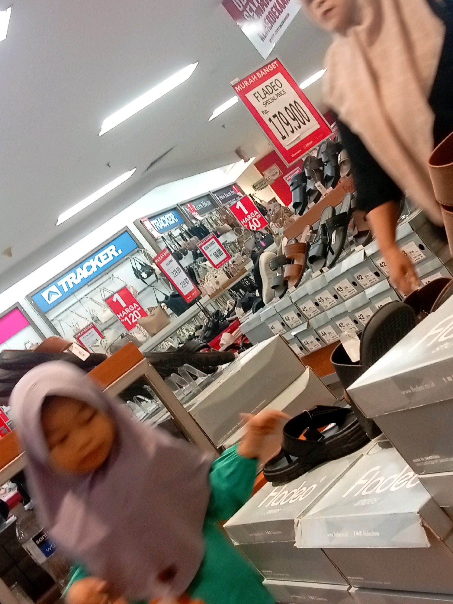 A dimly lit shoe store in Batam, Indonesia, features a ceiling with fluorescent lighting. The foreground is dominated by a young child wearing a light purple hijab and green shirt, holding an orange object. To the right, a woman in a light brown headscarf and dark clothing is partially visible. Behind them, shelves display numerous shoe boxes and shoes, with promotional signs visible, including one for "FLADEO" with a price of "179.900". Another sign reads "TRACKER." The environment appears to be a retail setting, possibly a department store or a dedicated shoe shop, with stacked boxes indicating a retail display.