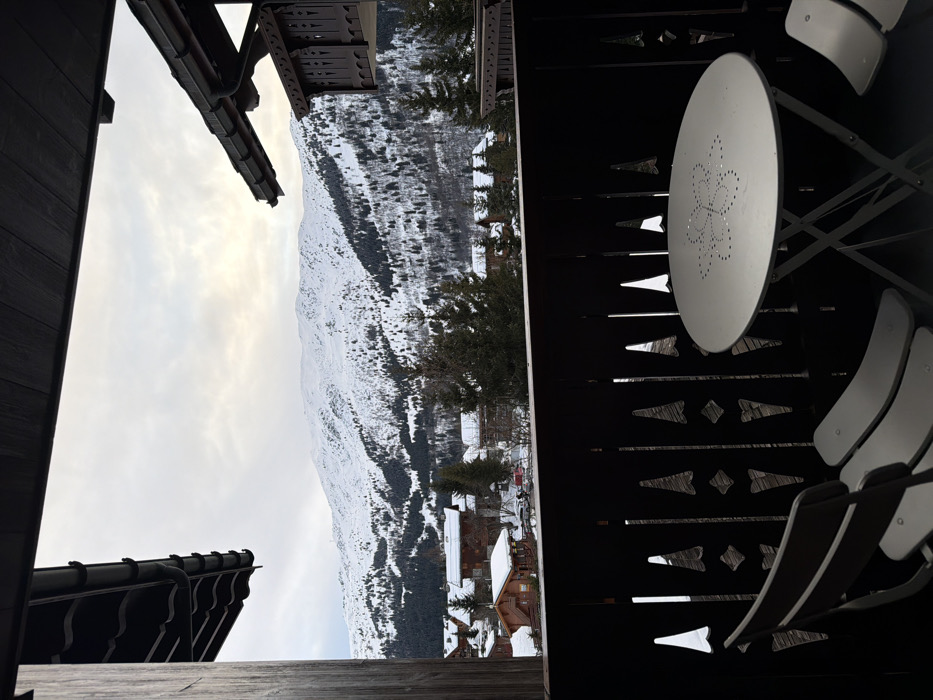 A balcony view showcases a snowy mountain landscape with scattered trees and buildings. A white round table and matching chairs occupy the right side, framed by a dark, decorative balcony railing. Roof elements of buildings appear on the left, while a cloudy sky fills the upper left corner.
