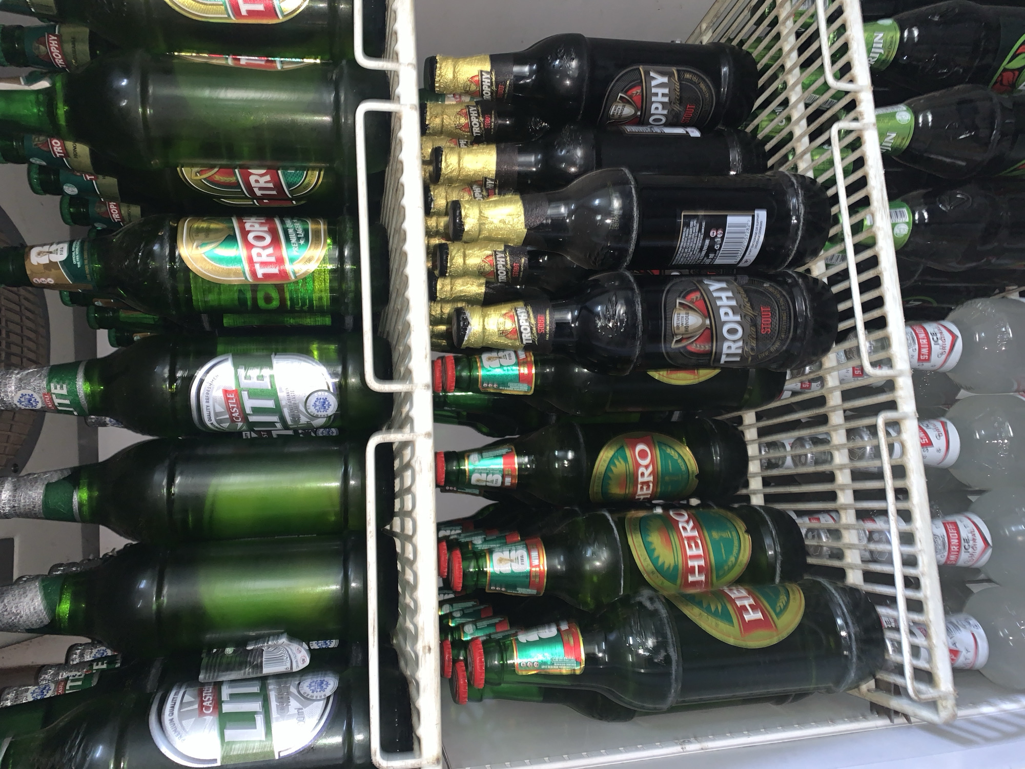 The image depicts the interior of a refrigerator filled with an assortment of bottled beverages, predominantly beer. The refrigerator is stocked on multiple shelves. The top shelf displays several green glass bottles, some with silver caps and labels indicating "CASTLE LITE". Other bottles on this shelf are less visible but appear to be similar in type.
The middle shelf is also packed with bottles. Some are green glass bottles with labels featuring gold and red accents, some with "TROPHY" written on them. To the right, there are darker bottles, possibly also beer, with gold foil caps and labels, some of which read "TROPHY SPECIAL STOUT". There are also some green bottles with red caps and labels.
The bottom shelf contains a mix of items. Some are clear glass bottles with white and red labels that read "SMIRNOFF ICE ORIGINAL". Interspersed among these are more green glass bottles, some with red caps and labels. The overall impression is a well-stocked beverage refrigerator, likely in a commercial setting or a very well-equipped home, given the variety and quantity of beverages. The location is identified as New Stadium, Nigeria.
