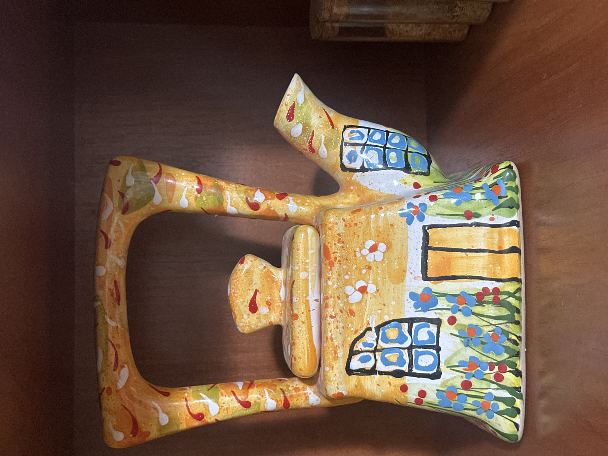 A whimsical, hand-painted ceramic teapot shaped like a cottage sits on a wooden shelf in what appears to be a display cabinet. The teapot is primarily yellow with white speckles and adorned with colorful designs, including red and green splatters, blue and orange flowers, and windows with blue panes. A brown wooden shelf supports the teapot, and the background is a dark wooden surface. To the right of the teapot, a blurred cylindrical object, possibly a jar, is visible. The scene is static and indoors. There is no indication of the city of Chornomorske, Ukraine, in the image or its context.
