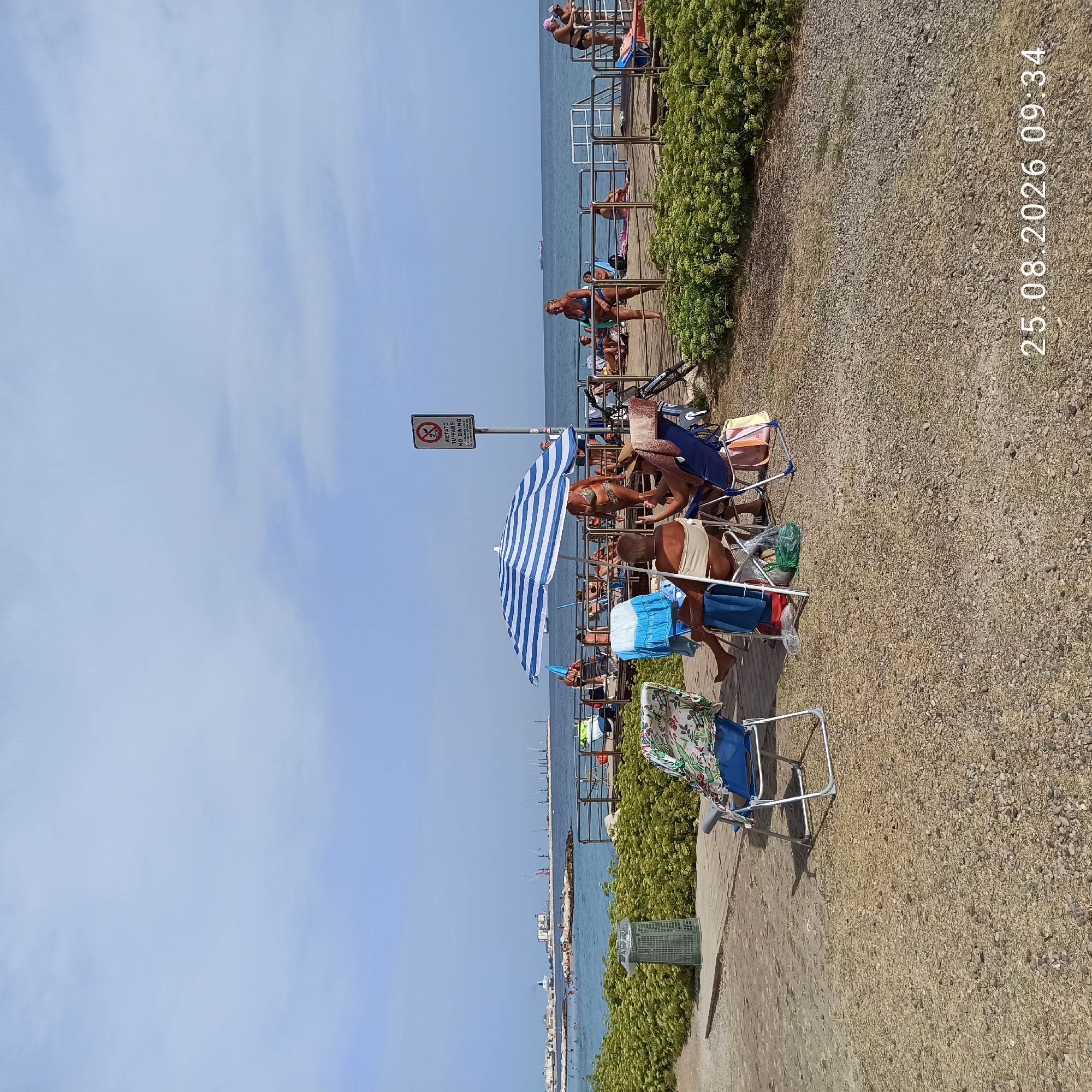 This image depicts a sunny day at a beach, possibly located in Bari, Italy, given the timestamp indicating August 25, 2026, at 09:34. The foreground features a gravelly ground area with several beach chairs, one of which is occupied by a person sitting and gesturing. Another beach chair is draped with a patterned fabric. To the left, a green trash bin is visible.
A blue and white striped umbrella shades a sitting area where two people are present. One person is seated, and another is standing. Further back, a wooden walkway is lined with metal railings. Beyond the walkway, people are seen sunbathing and swimming in the ocean. In the distance, the coastline features buildings and several construction cranes. A ship is visible on the horizon. The sky is mostly clear with some light clouds. A sign is posted on a pole, with Italian text that includes "VIETATO TUFFARSI NO DIVING".