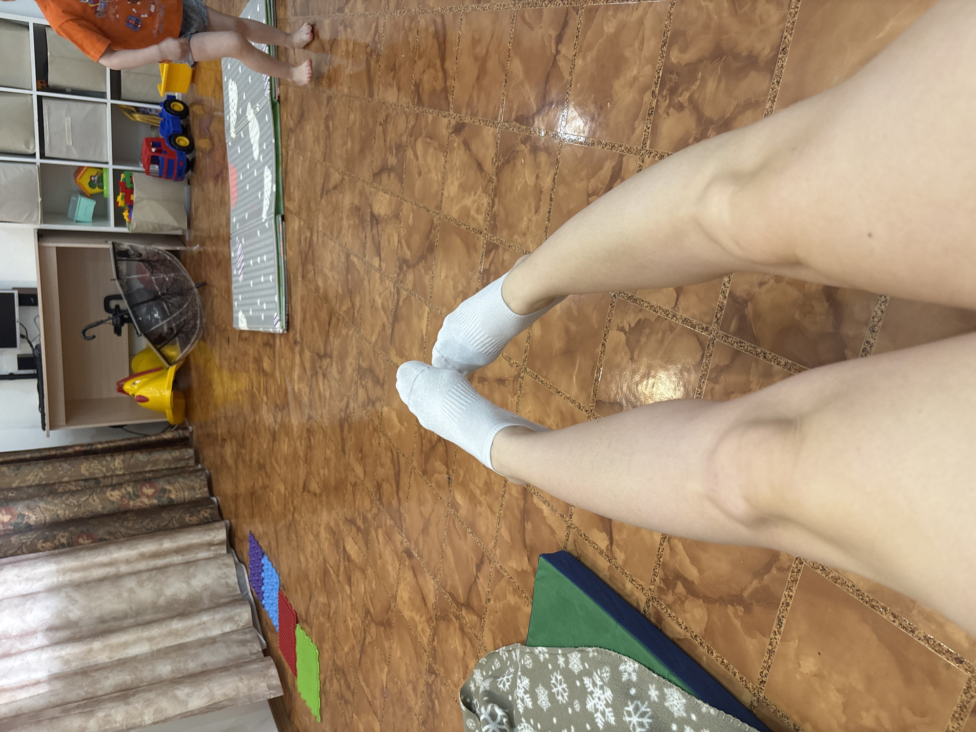 The photograph shows a first-person view of a person's legs in white socks on a tiled floor. In the background, a child is visible standing near a play mat and toy storage unit. The location may be Ферма, Russia, but there is no specific evidence in the image to confirm this claim.