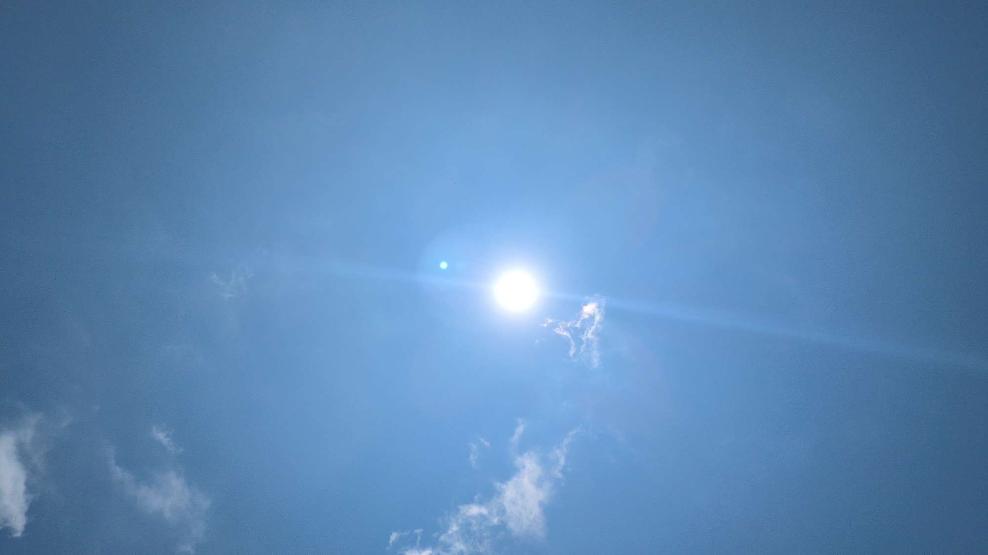 The image shows a bright, sunlit blue sky with a few scattered white clouds. The sun is prominently positioned in the center of the frame, creating lens flare. No people, subjects, objects, actions, or interactions are visible. The environment suggests an outdoor setting with clear weather. The city of Phuoc Dinh, Vietnam cannot be determined from the image.