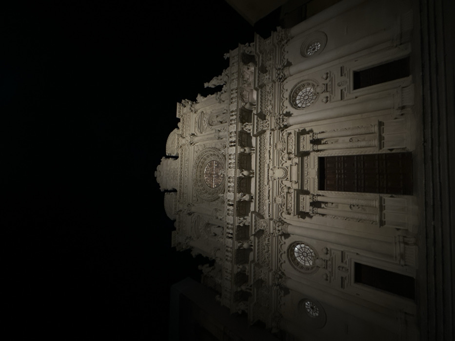 A low-angle, nighttime photograph captures the illuminated Baroque facade of the Basilica di Santa Croce in Lecce, Italy. The prominent structure, constructed of light-colored stone, dominates the frame against a black, featureless sky. The facade is richly detailed with extensive sculptural ornamentation across multiple tiers. A large, dark wooden main portal is centered at the base, flanked by two smaller, darker recessed doorways. Above the main portal, a large rose window with intricate tracery is visible. Two smaller circular windows are positioned above the side doorways. The facade features numerous columns, pilasters, pediments, and niches containing statues and figures, including a balustrade adorned with human and animal figures on the upper tier. Steps leading up to the church entrance are visible in the immediate foreground. No people or moving objects are present. Artificial lighting accentuates the architectural details and casts subtle shadows.