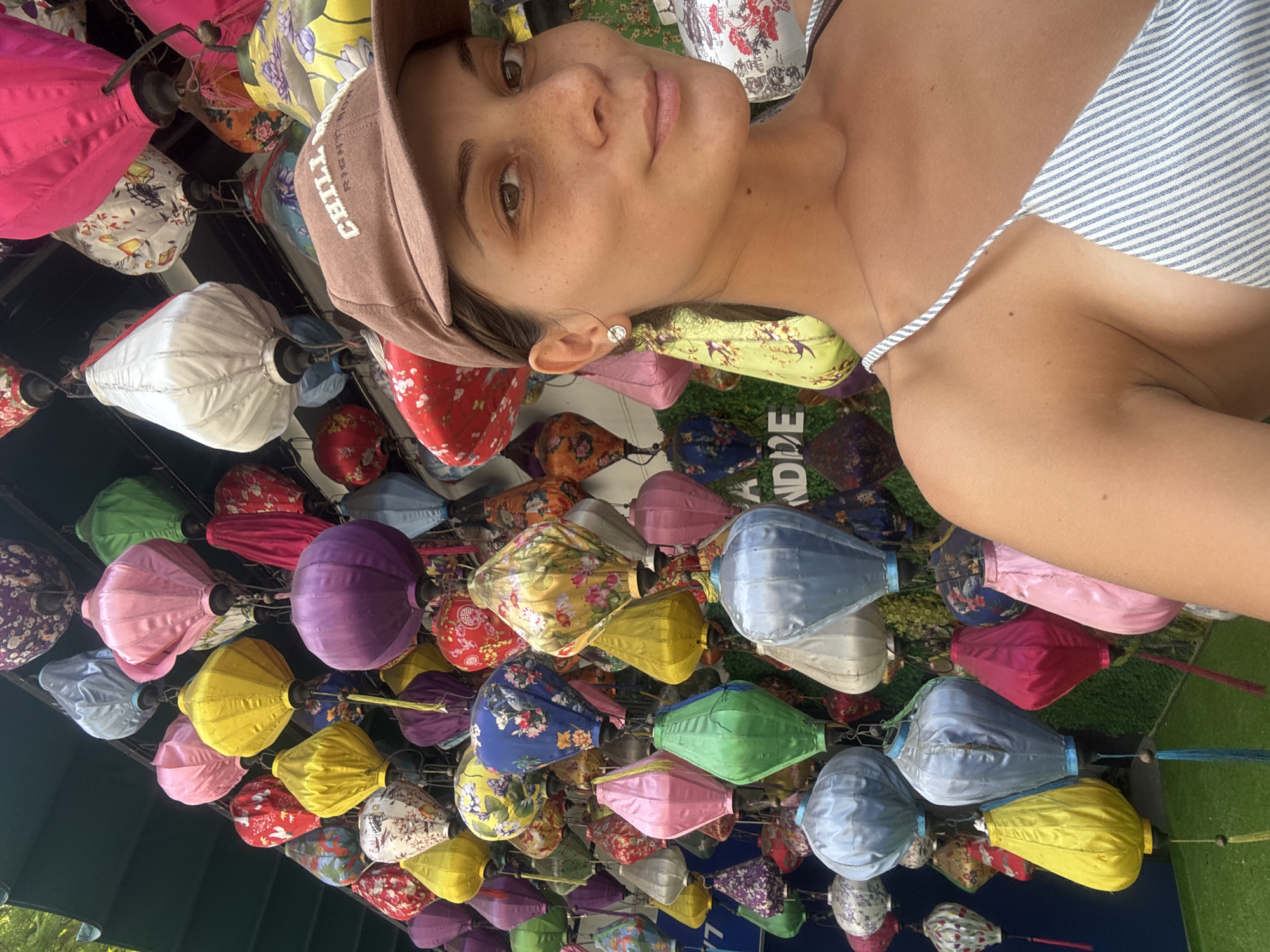 A selfie shot of a young woman with light skin, a brown baseball cap, and a striped top, appears to have been taken from below the colorful lanterns hanging from the ceiling. The lanterns are varied in color and design, predominantly in hues of red, pink, purple, blue, yellow, and green. Many of the lanterns have intricate floral or patterned designs on them. The context suggests this may be a tourist location in Đà Nẵng, Vietnam.