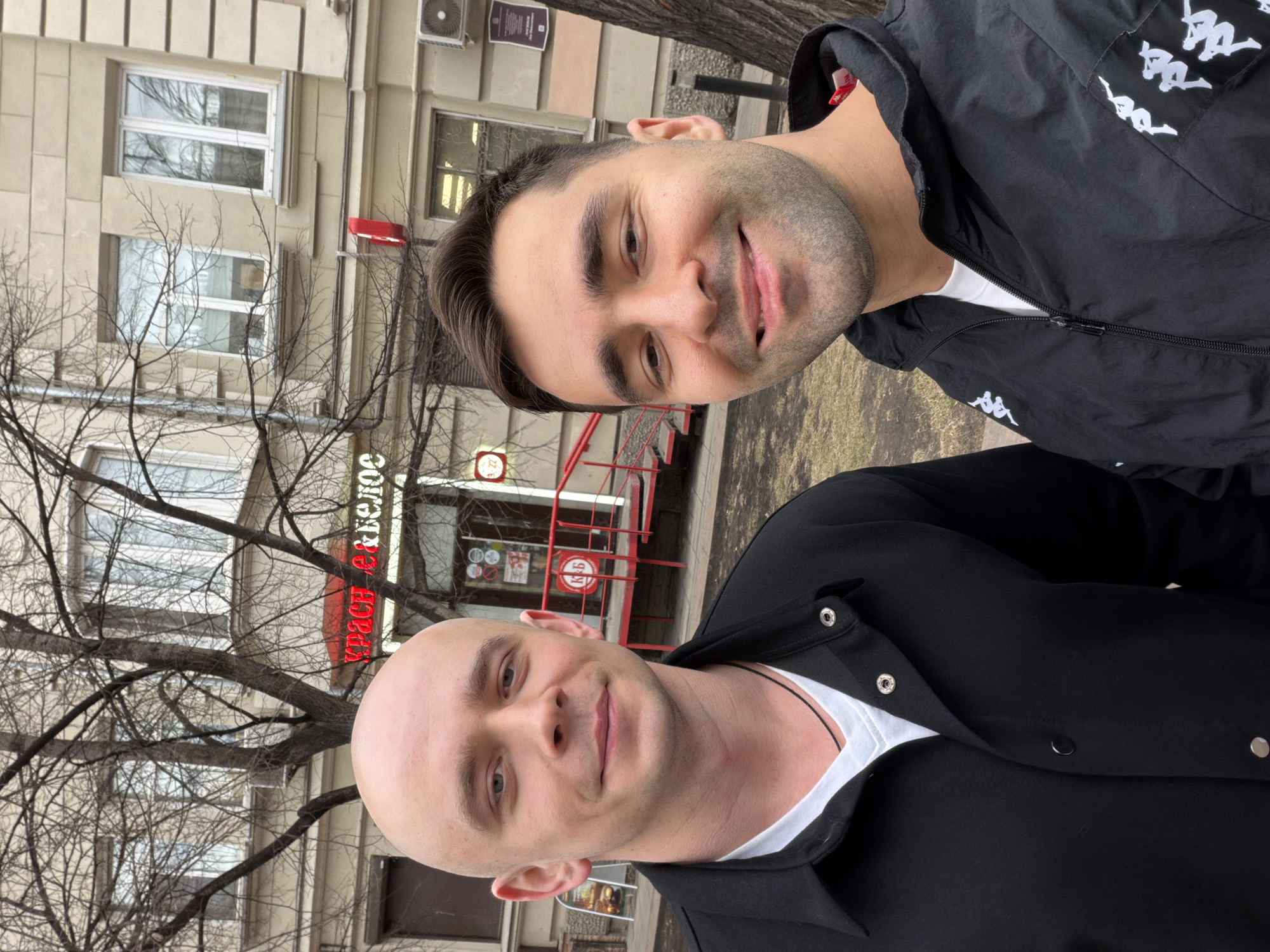 Two men are captured in an outdoor portrait, rotated 90 degrees clockwise. The setting appears to be an urban environment, likely Krasnoyarsk, Russia, as per the provided context.
In the foreground, a bald man with a light complexion and blue eyes is looking towards the camera with a subtle smile. He is wearing a black collared jacket over a white t-shirt, with a black cord or necklace visible under his collar.
Behind him, another man with short dark hair and stubble smiles broadly at the camera. He is dressed in a black hooded jacket featuring several white "Kappa" brand logos on the shoulder and chest area.
The background shows a light-colored building with multiple windows, some reflecting bare tree branches. These leafless trees, along with the overall lighting, suggest daylight on an overcast day, indicative of a colder season such as late autumn, winter, or early spring. An air conditioning unit is visible on the upper part of the building. A red metal railing and a red sign, partially obscured by branches and inverted due to the image rotation, display text. The sign reads "КРАСН & БЕЛОЕ" (Krasnoye & Beloye), a common store name in Russia, confirmed by a "К&Б" logo on the railing. Both men appear friendly and content.