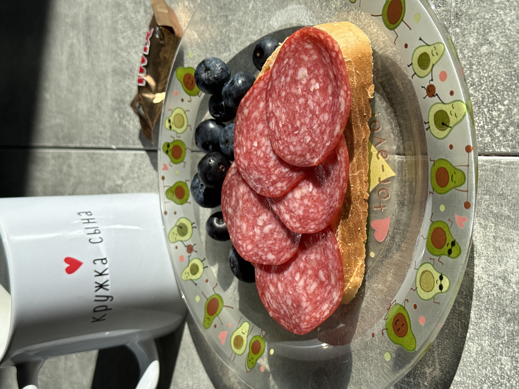 The image displays a breakfast setting on a grey tiled surface, likely a countertop or table, in Lyubertsy, Russia. In the foreground, a clear glass plate with a playful avocado pattern holds a slice of bread topped with several slices of red salami and a scattering of blueberries. Behind the plate, a white ceramic mug with a red heart and Russian text that reads "кружка сына" (son's mug) is visible. To the right of the mug, a partially unwrapped chocolate candy bar rests on the tiled surface. The lighting suggests a sunny day, casting distinct shadows.