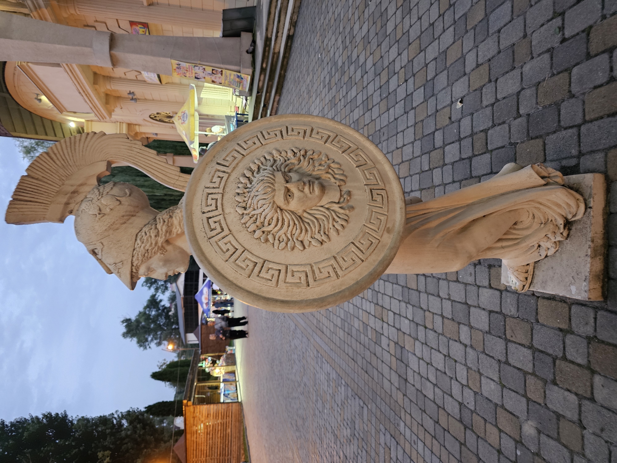 A statue of Athena, the Greek goddess of wisdom and warfare, stands on a cobblestone path in Cherkessk, Russia. The statue is made of light-colored stone and depicts Athena wearing a helmet and holding a shield with a Gorgon's head in relief. In the background, there are several wooden stalls and a building with columns. People can be seen walking in the distance. The sky is overcast, and it appears to be dusk.