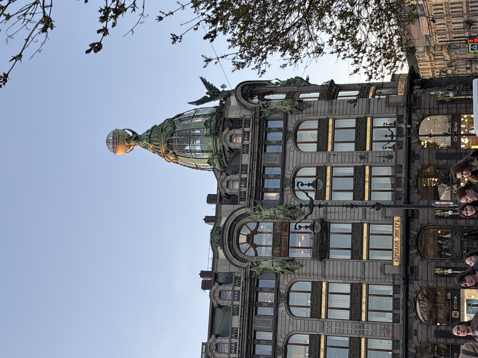 The image shows the Singer House building in Saint Petersburg, Russia. The building's architecture is prominent, with a glass dome topped by a sphere and sculptural details. The facade is ornate, and the building appears to be well-maintained. Bare tree branches are visible in the upper left and right of the frame. Part of a building with architectural details is seen on the right. A group of people is positioned on the right side of the building. The sky is clear.