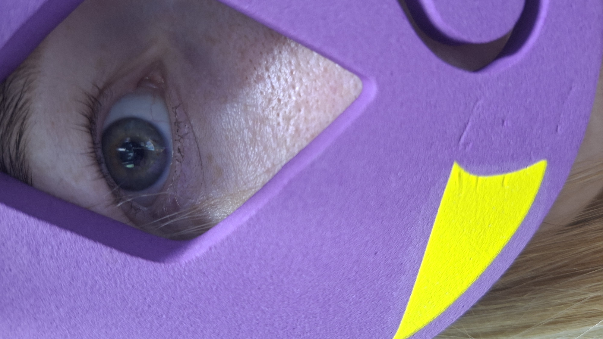 A close-up photograph depicts a single human eye peering through a purple mask with a yellow accent. The subject's skin is visible around the eye, exhibiting pores and fine hairs. The mask's material appears textured. The eye has an iris with variations in color. The setting is unknown, and no information connects the image to Chaikovsky, Russia.