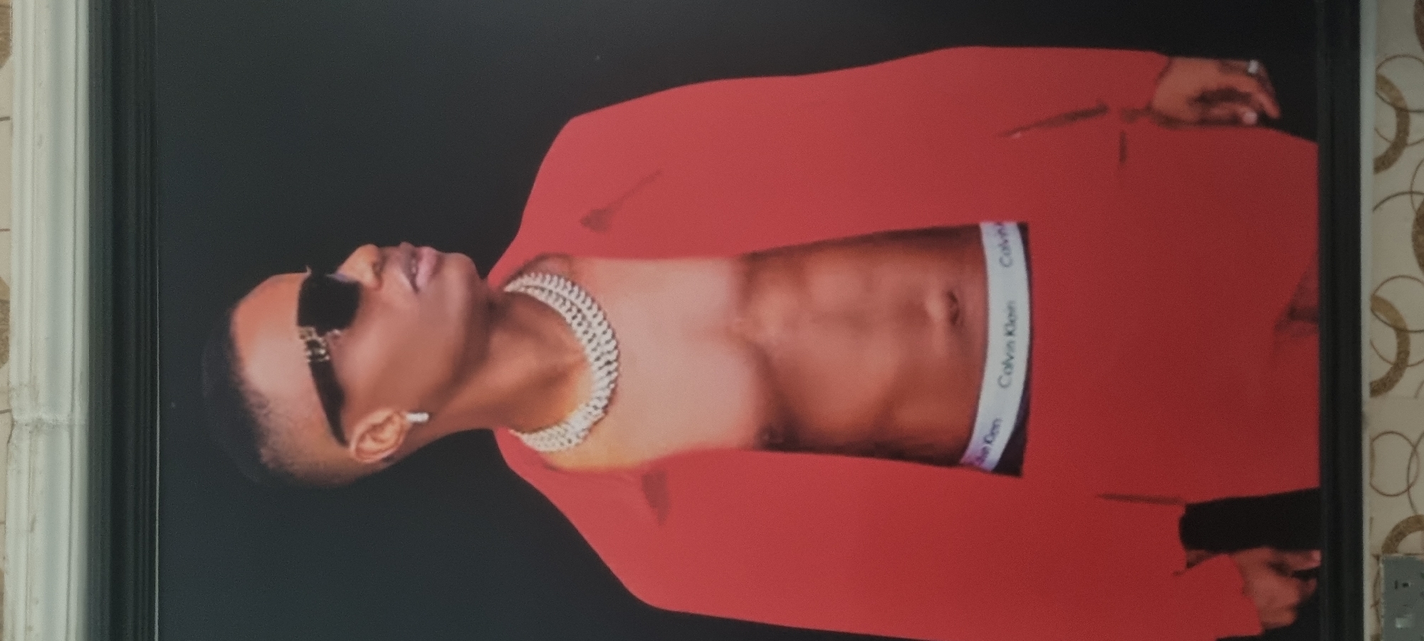 The image displays a head-to-mid-torso shot of a man, likely a celebrity, posing against a plain black background. The photograph, which appears to be a framed print, is currently rotated 90 degrees counter-clockwise.
The man, who has dark skin and short, neatly cut hair, is wearing dark sunglasses that obscure his eyes. He has a slightly open mouth, as if about to speak or in a relaxed pose. A prominent, multi-layered silver or white chain necklace adorns his neck. He is dressed in a vibrant red blazer or jacket, which is open, revealing his bare chest and abdomen. Below his navel, the waistband of a pair of boxer briefs is visible, clearly displaying the brand name "Calvin Klein" in white text on a dark band. His left hand, adorned with a ring on one finger, is slightly visible towards the right side of the frame.
The overall scene suggests a professional photoshoot or promotional image, emphasizing a stylish and confident demeanor. The lighting appears to be controlled studio lighting, highlighting the subject against the dark background. The context provided indicates this image is located in Alasia, Nigeria, although no specific environmental cues within the image itself confirm this.