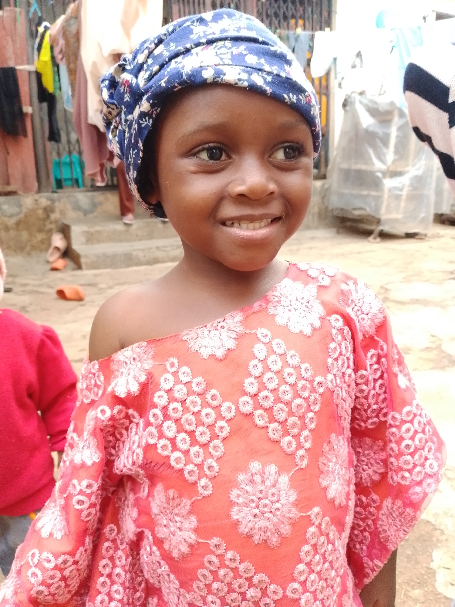 A young girl with dark skin and a blue and white floral headscarf smiles at the camera. She is wearing a pink off-the-shoulder dress with intricate white floral embroidery. In the background, laundry hangs on a line, and another person in a red shirt is visible to the left. The setting appears to be outdoors in a residential area.
