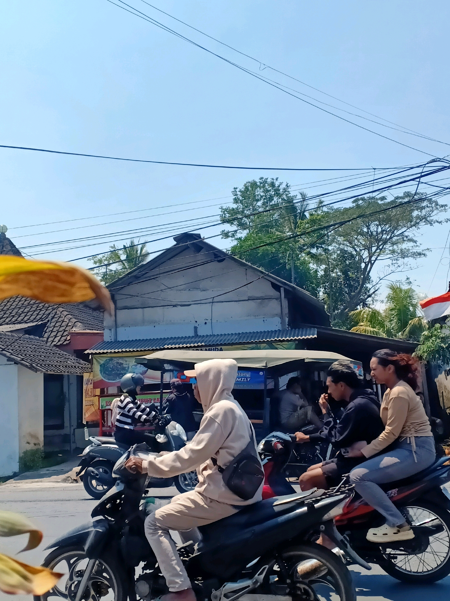 The image depicts a street scene in Jarit, Indonesia, on a bright, sunny day. In the foreground, two motorcycles are visible. The first, closer to the viewer, is black and is ridden by a person wearing a tan hooded sweatshirt and pants. They are looking forward, seemingly in motion. Behind this motorcycle, a red and black motorcycle is occupied by two individuals. The rider is a male with dark hair wearing a black shirt and shorts, while the passenger is a female with dark hair wearing a tan long-sleeved shirt and blue jeans. Both appear to be looking to the right. Further back, another motorcycle is seen with a rider wearing a black and white striped shirt and a helmet, and a passenger behind them.
The background reveals buildings with corrugated metal roofs, some with exposed beams. One building has a banner displaying text, possibly advertising food. Trees with lush green foliage are visible behind the buildings, and power lines crisscross the sky. A small red and white flag, likely the Indonesian flag, is visible on the right side of the image. The overall environment suggests a rural or semi-urban setting within Indonesia.