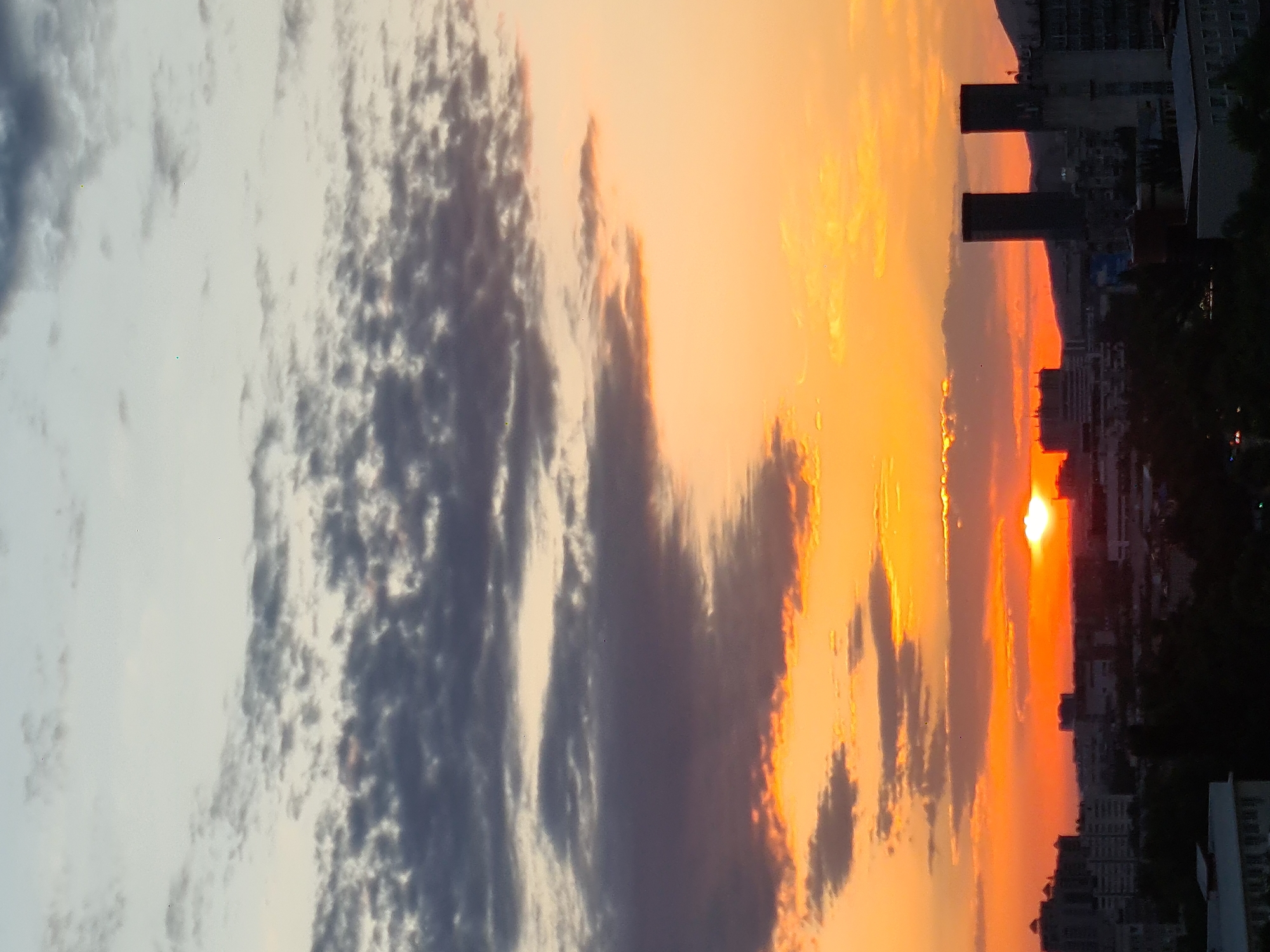 This is a vertical, eye-level photograph capturing a sunset over the city of Almaty, Kazakhstan. The sky is dominated by a gradient of colors, transitioning from pale blue and white at the top to a vibrant orange and yellow near the horizon. Fluffy, dark gray clouds are scattered across the sky, with some appearing as wispy streaks and others as more defined masses.
In the foreground, silhouetted against the sunset, is the Almaty cityscape. Buildings of varying heights are visible, including apartment blocks and what appear to be office buildings. Two prominent, dark, rectangular structures, possibly skyscrapers or towers, stand out in the mid-ground. In the far distance, a mountain range is faintly visible against the bright sky. The sun is partially obscured by clouds, creating a soft, glowing orb near the horizon. There are no discernible people or animals in the image. The overall atmosphere is one of a calm evening in an urban environment.