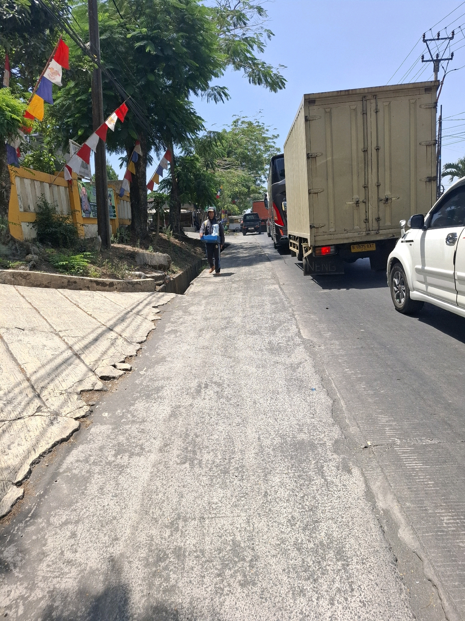 A street scene in Sukalarang, Indonesia, captured from a low angle. The foreground shows a damaged asphalt road with a concrete embankment to the left, exhibiting cracked and uneven surfaces. A person, identified as a woman by her attire, is walking away from the camera on the right side of the road, carrying a blue container. She is dressed in dark clothing, including a headscarf, and is wearing dark pants and shoes. In the background, traffic is visible, including a large beige cargo truck with license plate B 9875 BXT, a white car, and other vehicles partially obscured. Flags, likely Indonesian flags due to their red and white colors, are displayed on poles and strung between trees on the left side of the street. The environment appears to be a roadside with lush green trees and vegetation bordering the area. The sky is clear and blue, indicating a sunny day.