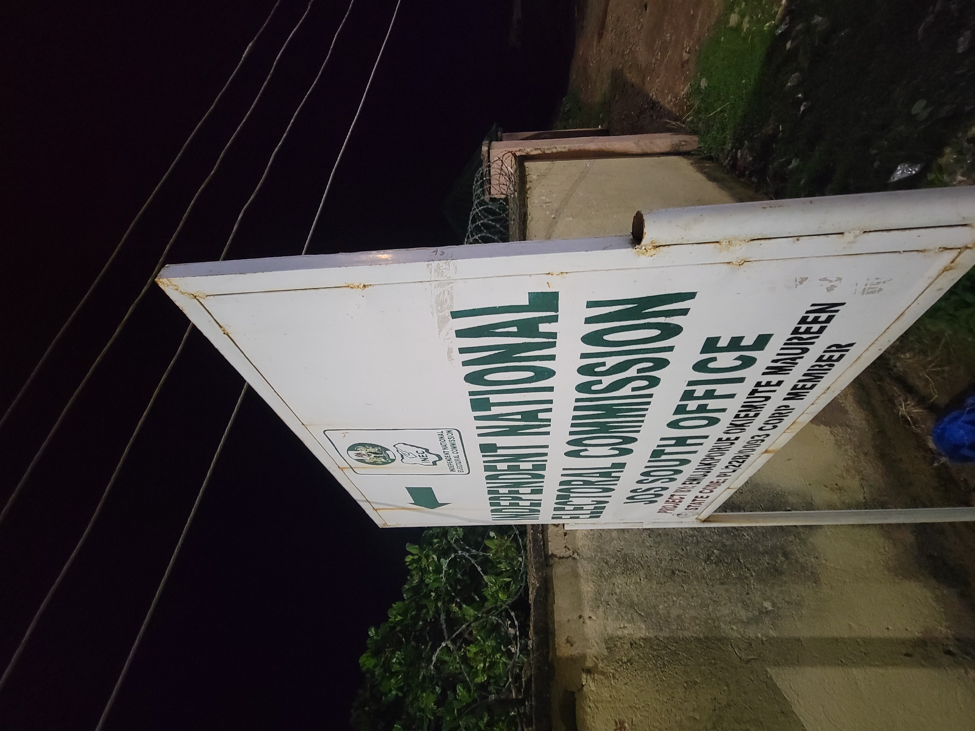 This is an outdoor nighttime shot of a sign for the Independent National Electoral Commission (INEC), specifically the JOS South Office. The sign is white with green lettering and features the INEC logo. A green arrow points to the right. The sign is positioned in front of a wall with a fence topped with barbed wire. A few tree branches and wires are also visible. The location is Unknown, Unknown.
