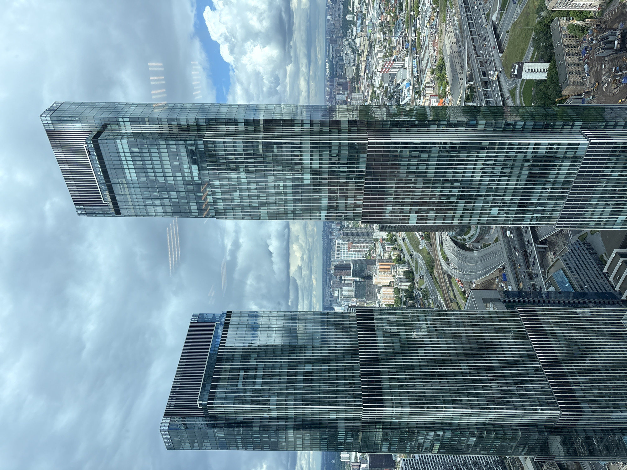 The image shows a high-angle view of two modern glass skyscrapers reflecting the sky. Below the skyscrapers, the cityscape stretches into the distance, featuring roads, buildings, and greenery. Khimki, Russia is not visible in the picture.