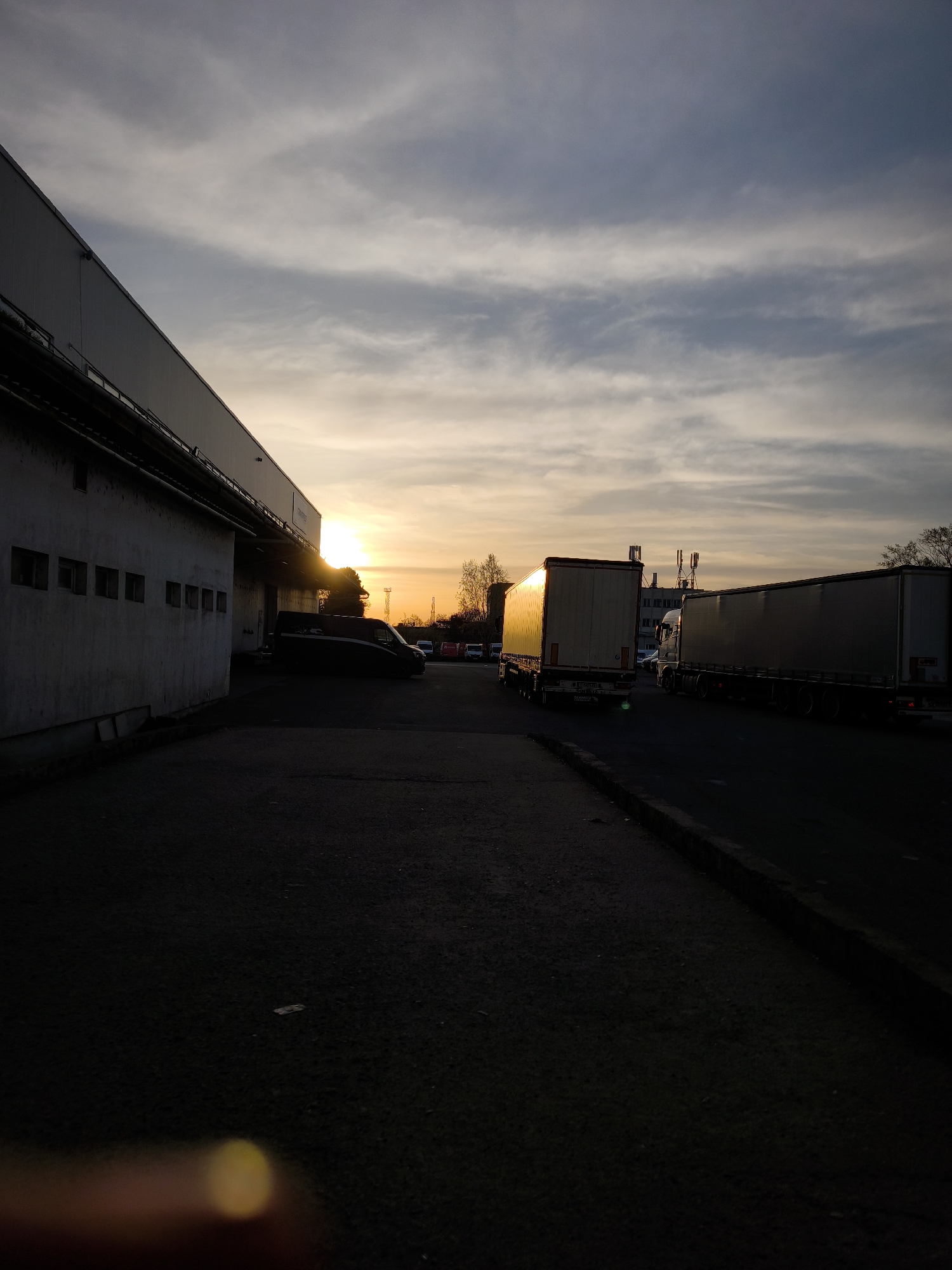 The image depicts an industrial scene at sunset in what appears to be a loading or parking area, with long-haul trucks parked alongside a building. The building is long, low, and possibly a warehouse or factory, with a white or light-colored exterior. The foreground is dark, showing the asphalt surface of the loading area.
The sun is setting on the horizon, providing a warm, golden glow to the scene. The sky is filled with streaky clouds that are illuminated by the setting sun, creating a striking contrast between light and shadow.
In the distance, there are other vehicles and structures, including what appear to be antennae or communications towers atop a building. There are also some trees with bare branches, suggesting it could be late autumn or winter.
The location is in an industrial area of Burgas, Bulgaria.