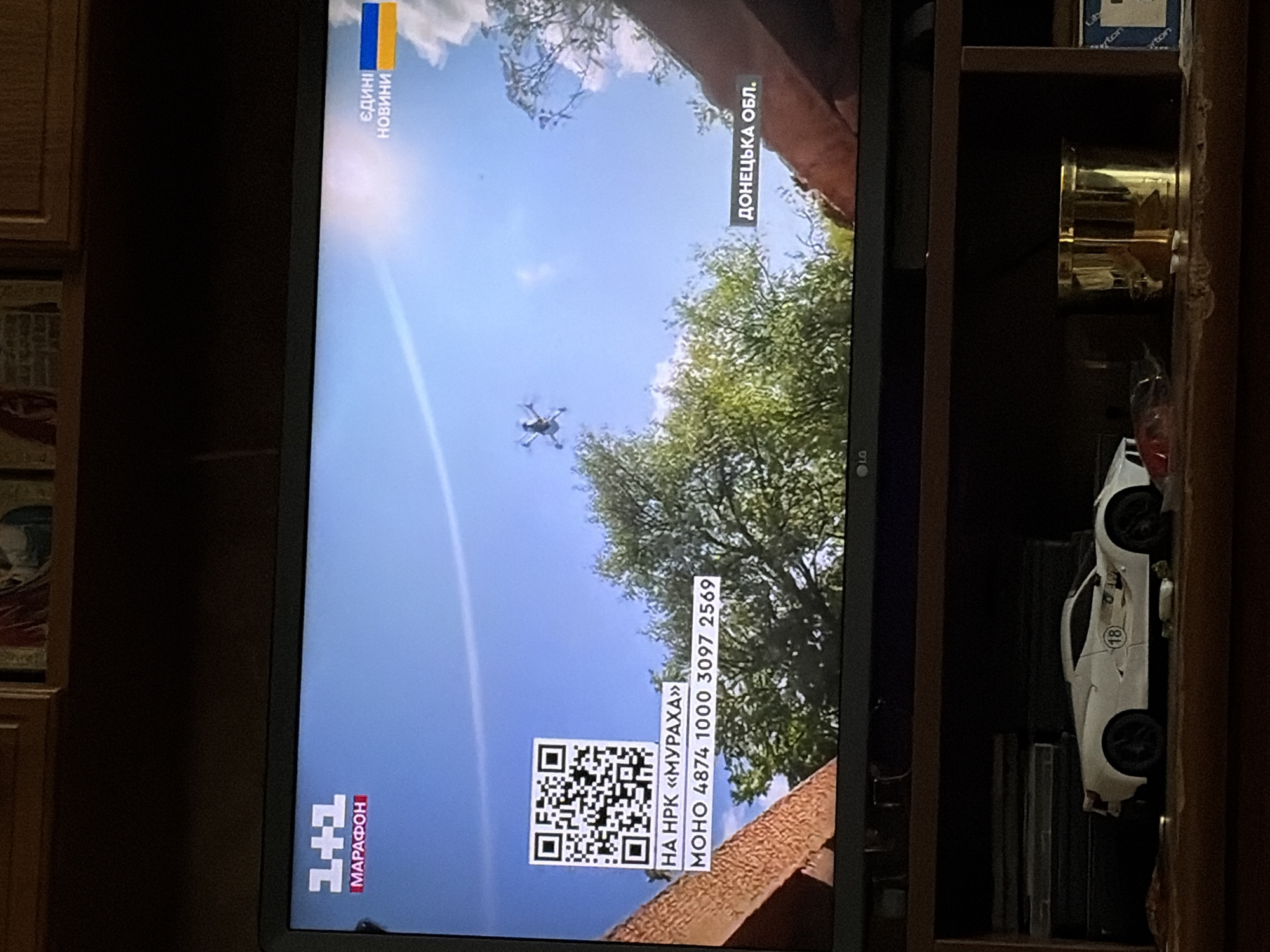 The image is a photograph of a television screen displaying a news broadcast. The screen shows a bright blue sky with scattered clouds and green trees. A drone is visible in the upper center of the screen, flying in the sky. In the lower left corner of the screen, there is a QR code and some text. In the lower right corner, there is text that says "ДОНЕЦЬКА ОБЛ." which translates to "DONETSK REGION".
The television itself is placed on a wooden shelf, below which are other objects. These include what appears to be a toy white sports car with the number "18" on the side, some stacked DVDs or video games, and a shiny gold cylindrical object. The background behind the television appears to be a wooden cabinet.
The image is taken from a slightly angled perspective, with the camera capturing the reflection of the room on the television screen.