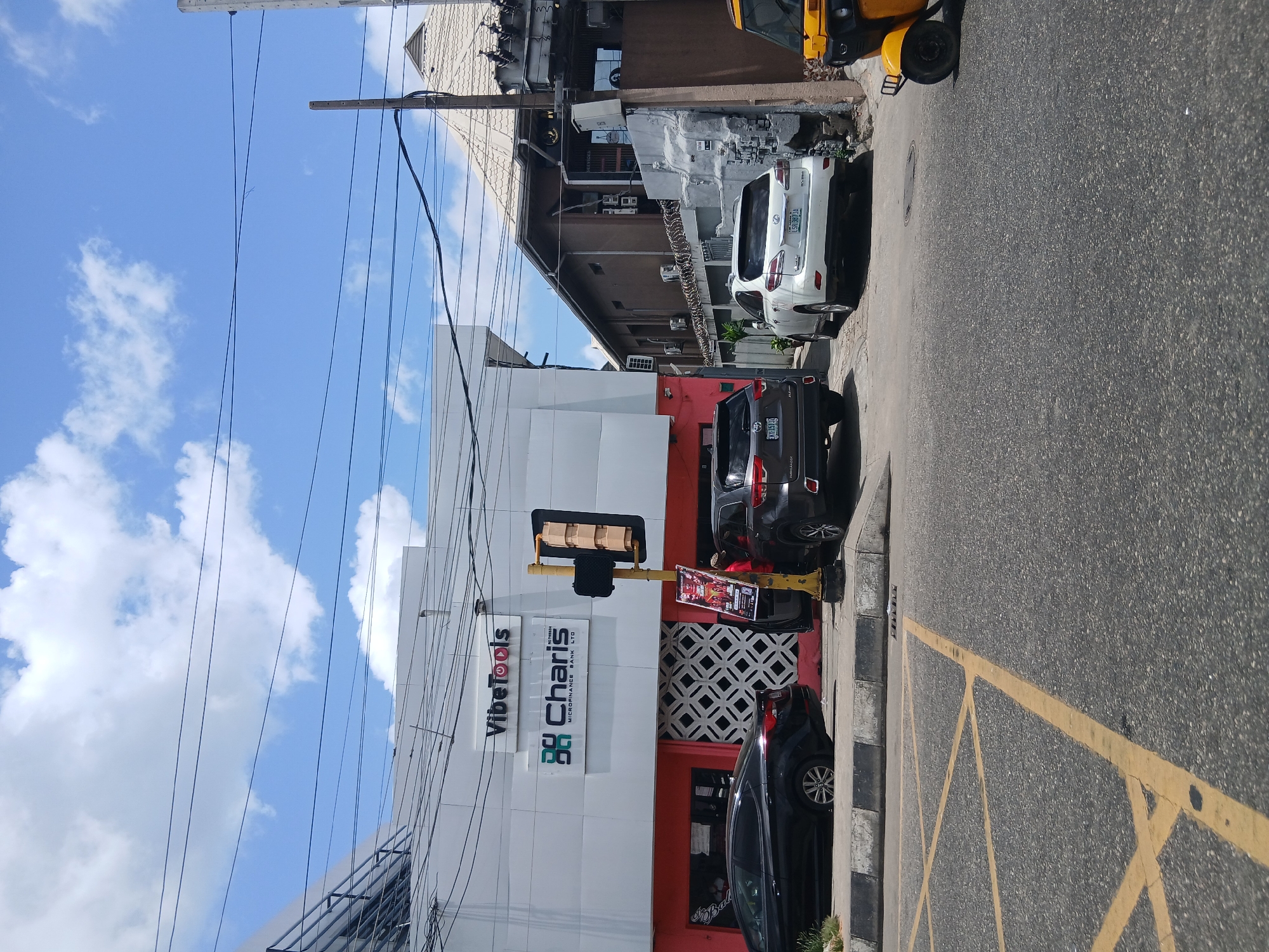 A bright daylight street scene in Animashawun, Nigeria, shows a commercial area with buildings, parked vehicles, and street infrastructure under a clear blue sky with scattered white clouds. The primary focus is a multi-story building on the left, featuring a white upper facade and a distinctive red lower section with geometric patterned vents.
Prominently displayed on the white part of this building are two signs. The upper sign reads "VibeTools" with a circular logo, and below it is "Charis Microfinance Bank LTD" with a square logo composed of interlocking 'C' shapes. On the red lower facade, partially visible text on a dark sign reads "RY BAL" or "OY BAL," potentially part of a larger business name.
Parked in front of this building are several vehicles. A black SUV, possibly bearing an "OYO" state license plate, and a black sedan are visible in the foreground. Further down the street, a white SUV with the Nigerian license plate "LSG 387 JA" is parked. Part of a yellow tricycle (known as a keke napep) is visible on the far right, suggesting local transportation.
A yellow traffic light pole with multiple signal heads stands on the sidewalk in front of the Charis building, and a small poster with red and black text is attached to its base. The asphalt road in the foreground features distinct yellow diagonal criss-cross markings, indicating a 'no parking' or 'keep clear' zone. Overhead, numerous power lines crisscross the sky, connecting to utility poles. In the background, behind the main white and red building, an older, darker brown building with what appears to be barbed or razor wire atop a wall is visible. The bright sunlight suggests midday.