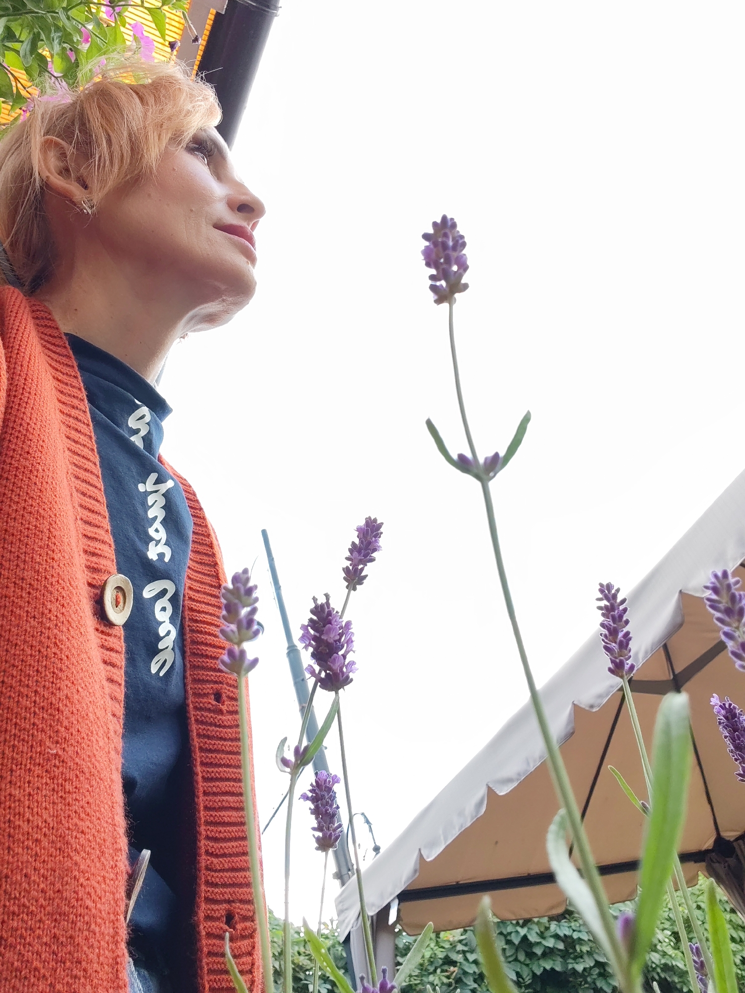 An adult female with short, light-colored hair, wearing an orange knitted cardigan over a dark blue shirt with white cursive writing, is looking upwards and to the right. She has light skin and is wearing earrings. She is positioned in the foreground, with several lavender plants and stalks in bloom in front of her. Behind her, a beige canopy with a scalloped edge is visible, with green foliage and possibly a fence or wall further in the background. The sky is overcast and bright. The location is identified as Bol'shie Mkhi, Russia.