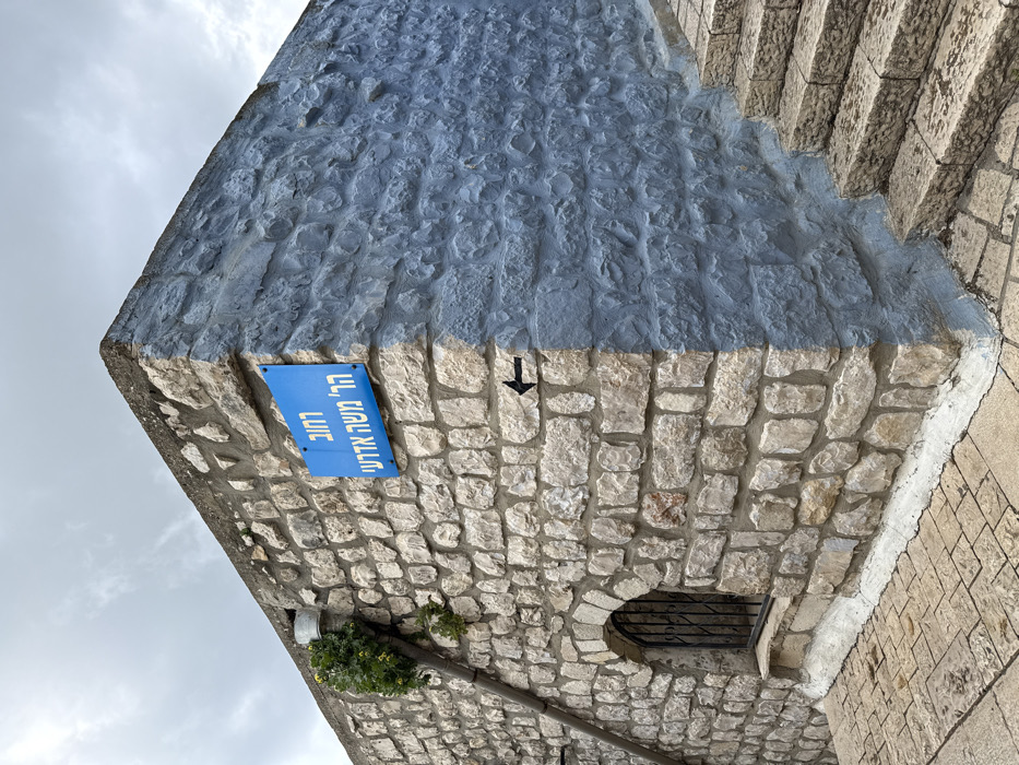 In the city of Safed, Israel, an outdoor scene captures the corner of a building at Harav Moshe Adre'i Street. The left facade of the building is constructed from irregular, light-colored stone blocks, while the right facade, turning the corner, is composed of similar stone blocks uniformly painted a medium-blue color. A sharp vertical line marks the transition between the unpainted and blue-painted sections at the building's corner.
A rectangular blue street sign with white Hebrew text, reading "רחוב הר' משה אדרעי" (Harav Moshe Adre'i Street), is affixed to the upper left portion of the light-colored stone wall. Below and slightly to the right of this sign, a small black arrow is painted directly onto the light-colored stone, pointing to the left. In the lower left section of the light-colored wall, an arched, recessed window opening is visible, secured by a black metal grate. A dark-colored downspout descends diagonally along the upper left area of the light-colored wall; a green plant with small yellow flowers grows around its upper section. To the right of the blue-painted wall, a series of worn, light-colored stone steps ascends out of view. The ground in the foreground is paved with irregularly shaped light-colored flagstones. The sky above is overcast, showing diffuse cloud cover.