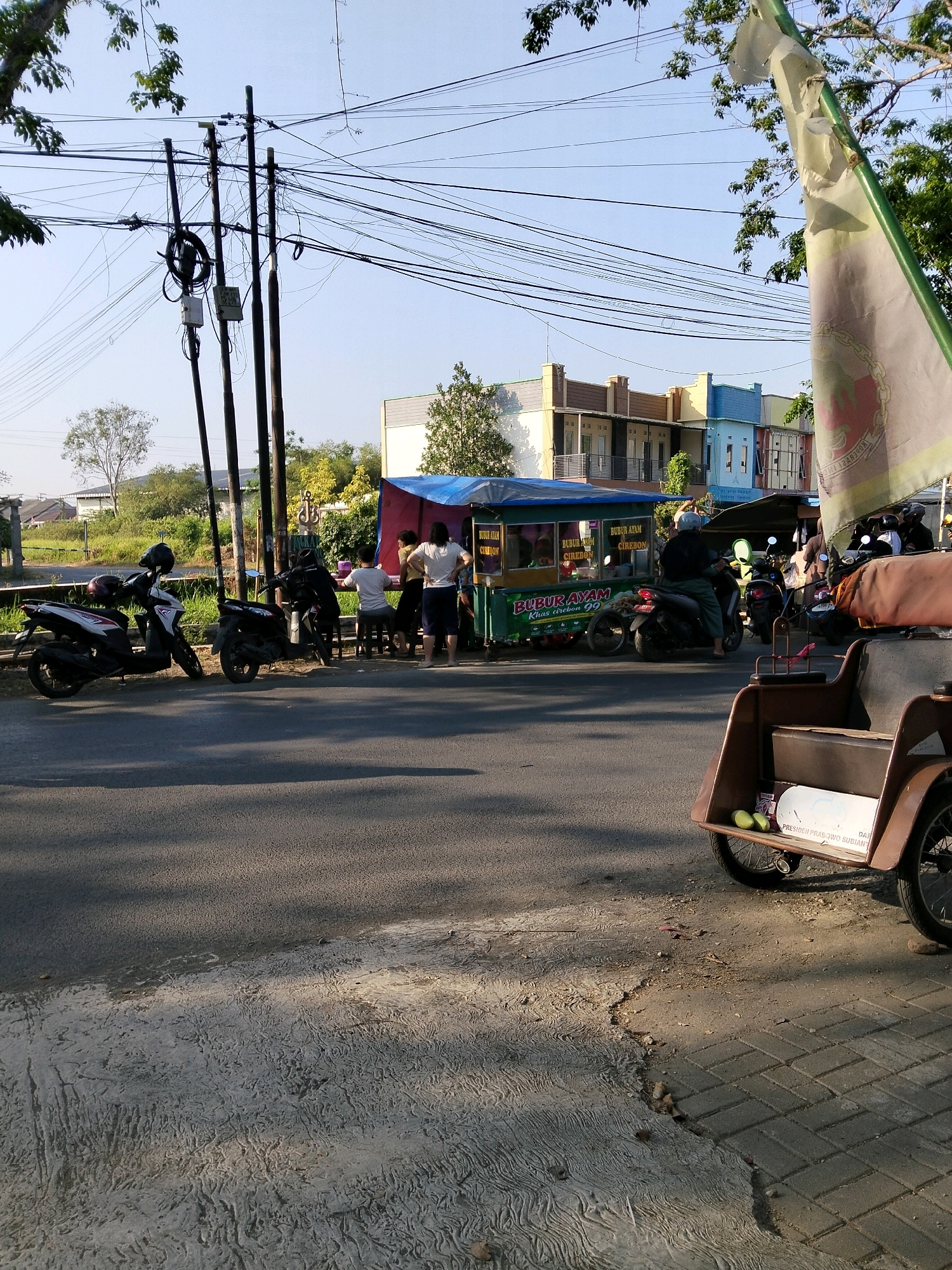 This is an outdoor, eye-level, medium shot of a street scene in Karangmalang, Indonesia. The image captures a roadside food stall with customers, several parked motorcycles, and a pedicab in the foreground.
The food stall is set up with a green and blue tarp for shelter. There are several plastic chairs and small tables around it. The stall appears to be selling "Bubur Ayam" (chicken porridge), as indicated by signage on the stall itself. A few people are standing or sitting around the stall, some appearing to be customers and one person on a motorcycle.
Parked to the left of the stall are two scooters. To the right of the stall, a brown pedicab is prominently featured in the foreground, facing towards the right of the frame. It has a white box on its side with text that appears to be a political slogan. Two green limes are visible on top of this box.
The background shows a clear blue sky with some overhead power lines and utility poles. Behind the stall, there is a row of colorful buildings, suggesting a commercial or residential area. Trees and vegetation are also visible in the mid-ground. The road surface is asphalt, with a concrete sidewalk or textured pavement in the foreground. The lighting suggests it is late afternoon, with long shadows cast across the road. The overall impression is a bustling street corner in a typical Indonesian town.
