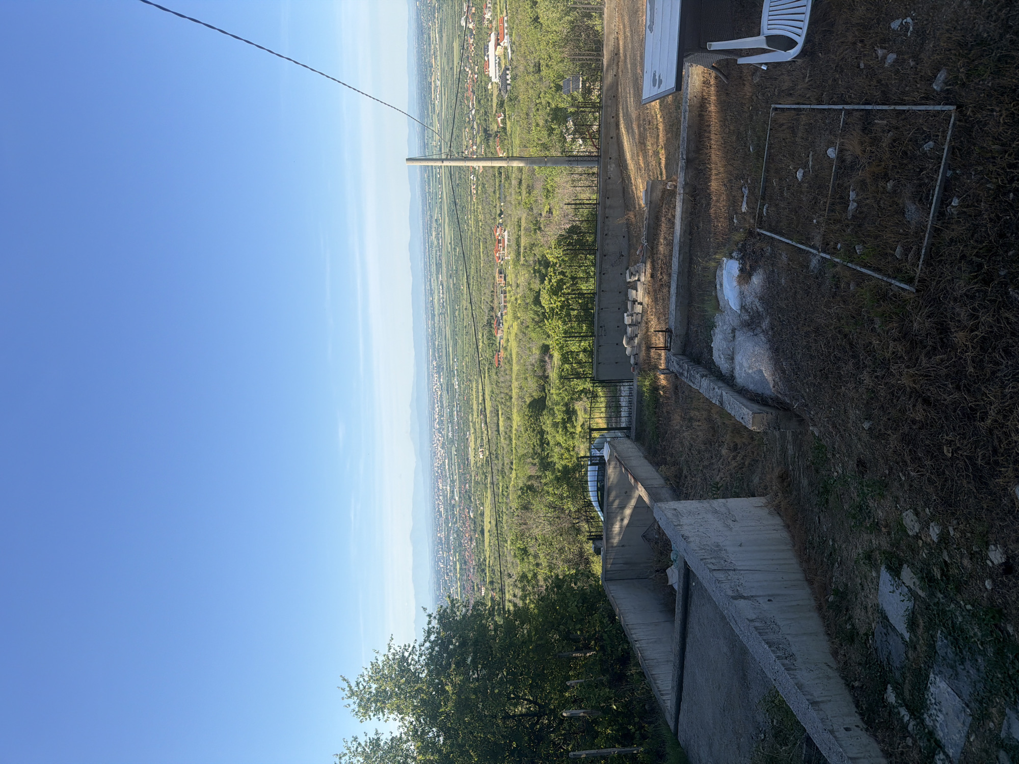 A landscape photograph taken from an elevated vantage point, likely in or near the city of Brestnik, Bulgaria, depicts a panoramic view spanning from a clear blue sky to a distant cityscape nestled amidst lush green vegetation. A utility pole with overhead wires stands prominently in the middle ground, visually connecting the foreground elements with the sprawling landscape below. The foreground features a concrete structure or retaining wall along with overgrown vegetation, suggesting an abandoned or undeveloped area. A metal frame or enclosure lies on the ground amidst dried grass, partially obscured by debris. A small white plastic chair sits near a flat white surface, possibly a section of a building or a large slab of concrete. The composition suggests a scene of observation or overview, capturing a blend of urban development and natural environment.
