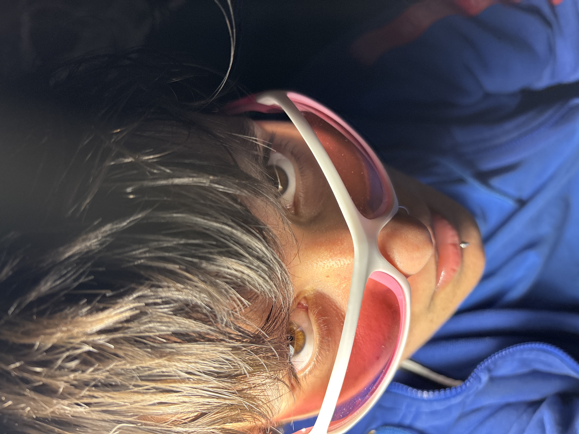 A close-up, tilted shot shows a person with tan skin, brown eyes, and short, dark brown hair wearing pink-tinted sunglasses with a white frame. The person has a lip piercing. The subject is wearing a blue garment with a zipper. The background is out of focus and dark. There is no indication of the location being Sofia, Bulgaria.