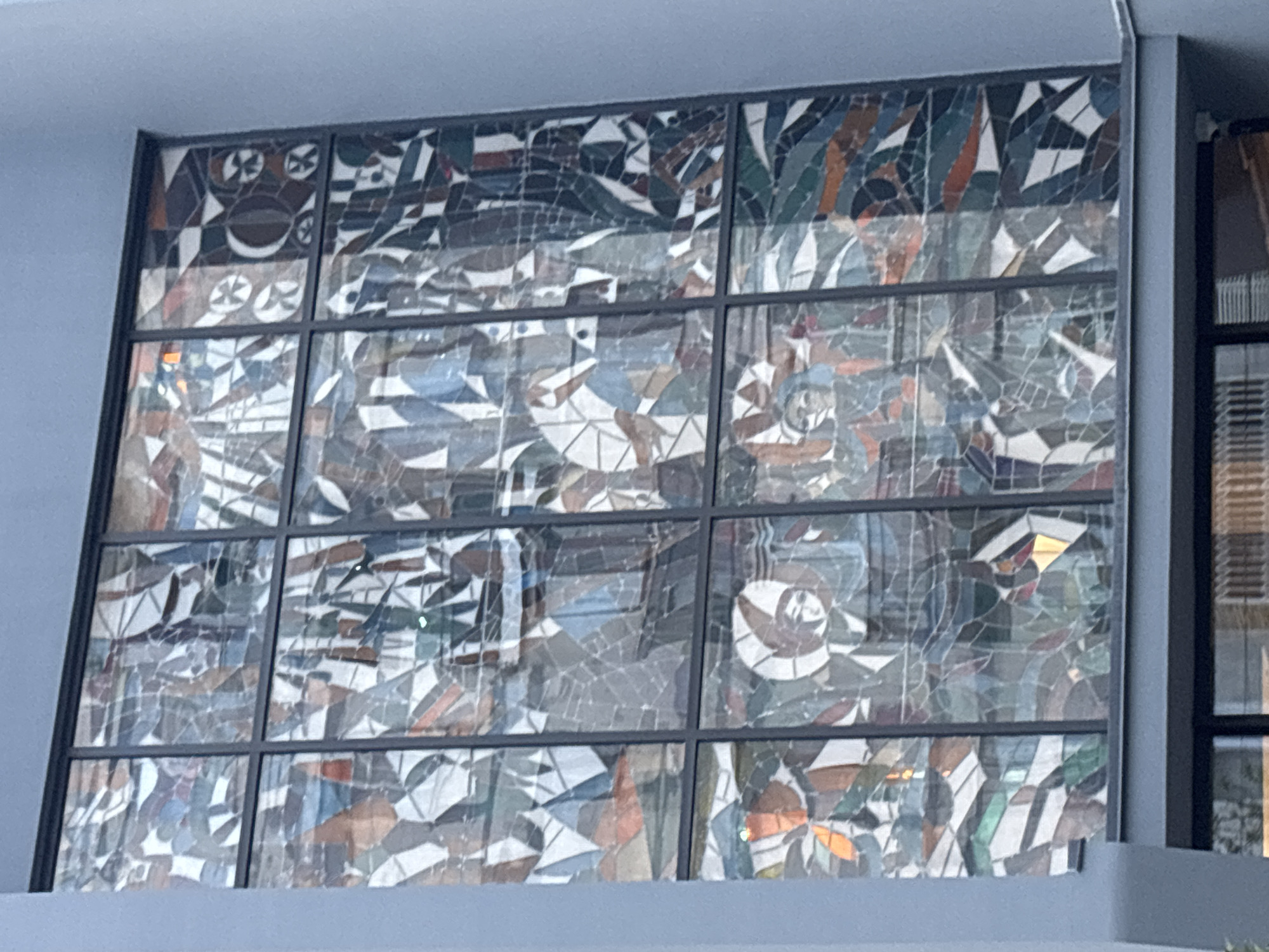A large, ornate stained glass window is the central subject of this exterior shot of a building in Tbilisi, Georgia. The window is framed by a light grey/blue building facade on the top, left, and bottom, with a darker vertical element and another window visible on the far right.
The stained glass itself is divided into a grid of panels by dark metal mullions, creating a mosaic of abstract and geometric shapes. The color palette primarily consists of various shades of blue, green, grey, brown, and white, accented with occasional warmer tones like orange and amber, particularly in the lower and right sections.
Within the intricate patterns of shattered-like glass pieces, several stylized human-like figures or motifs can be observed. In the upper left panel, a distinctive design resembling a stylized face or mask, with circular "eyes" and a curved "mouth," is flanked by smaller, circular, propeller-like emblems. Other sections of the window, especially towards the right and bottom, feature shapes that suggest human heads or busts, some adorned with halo-like circles or distinct head coverings, all integrated into the abstract design.
The lighting appears to be natural daylight, possibly indirect or overcast, as there are no strong shadows, and the colors of the glass are subtly diffused rather than brightly lit. The image captures a static architectural detail; no activity is taking place. No visible text is present on the window or the building facade. The window serves as a prominent artistic and architectural feature of the building.