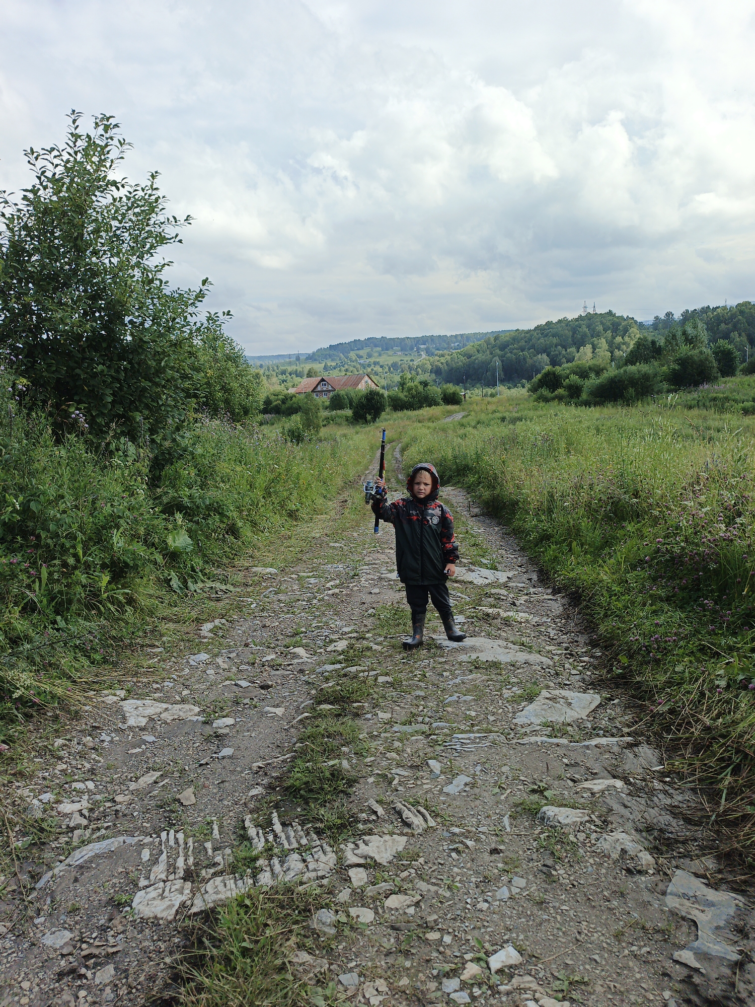In Medvedyovka, Russia, a young child stands on a dirt and gravel path, holding a fishing rod. The child is wearing a hooded jacket and rubber boots. The path is surrounded by green grass and trees, with houses and hills visible in the background under a cloudy sky.