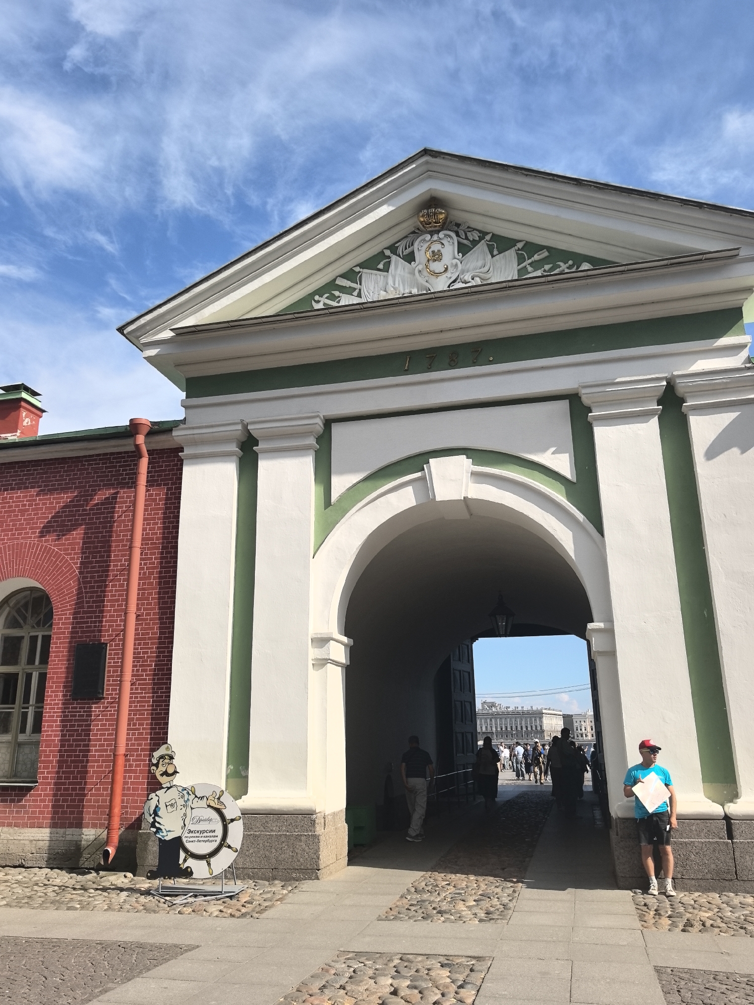 The image depicts an architectural facade featuring a large arched gateway. The building is painted in white and green, with a red brick section on the left. Above the arch, there is a triangular pediment adorned with decorative elements, including a coat of arms and the inscription "1787". A red drainpipe runs down the side of the brick building.
In front of the arch, on the left, is a cutout of a man dressed as a chef, holding a ship's wheel. This object appears to be a sign for tours or excursions, with Russian text visible on the wheel: "Экскурсии" (Excursions) and "Санкт-Петербург" (Saint Petersburg).
Through the archway, a street scene is visible. Several people are walking or standing, and in the distance, there are buildings characteristic of a European city. On the right side of the arch, a young person wearing a blue t-shirt, black shorts, a red cap, and sunglasses stands holding a white piece of paper.
The ground is paved with cobblestones. The sky is blue with scattered white clouds, suggesting it is daytime and the weather is fair. The overall impression is that of a historical landmark in Saint Petersburg, Russia, possibly an entrance to a fortress or an important historical site, with an element of local tourism promotion.