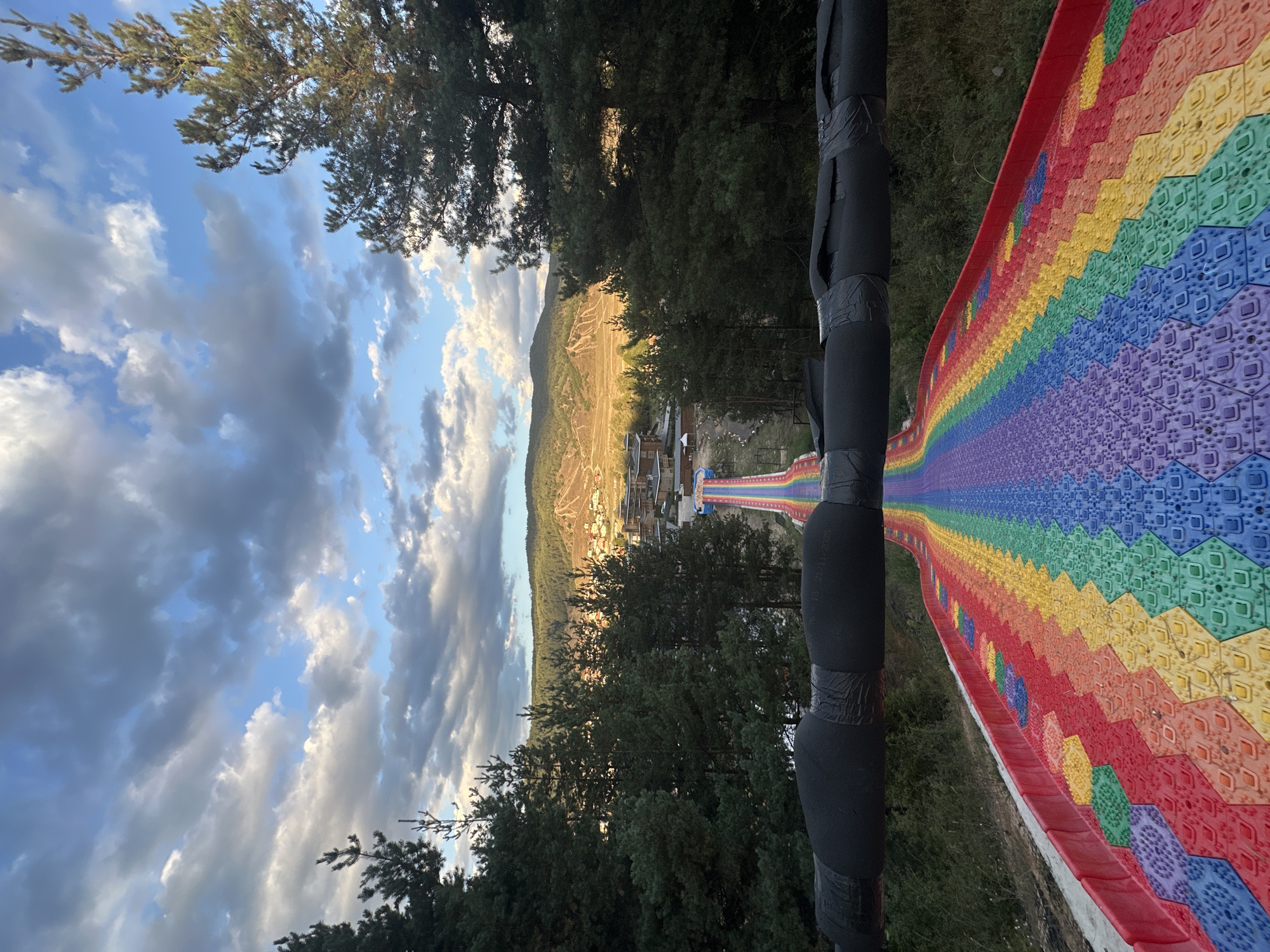 The image shows a long, rainbow-colored slide winding down a grassy slope. The slide is made of hexagonal tiles, with each color of the rainbow represented. The slide is surrounded by trees and in the background, there is a village with buildings and hills. The sky is cloudy with patches of blue. The scene appears to be taken from the top of the slide, looking down. This is the rainbow slide in Bania, Bulgaria, a popular tourist attraction.