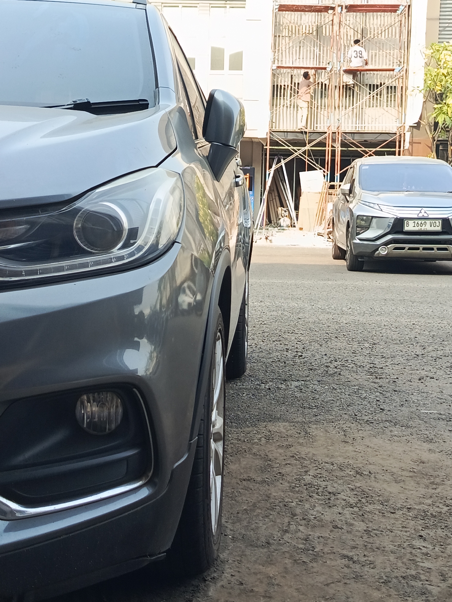 The image captures a street scene in Tangerang, Indonesia. In the foreground, the left side of a dark gray SUV is prominently visible, showing its headlight and bumper. To the right, further down the street, another gray SUV is parked, displaying a license plate that reads "B 1669 VOJ." Behind the vehicles, a building under construction is partially visible, with scaffolding erected on its facade. Two individuals are working on the scaffolding. One person is positioned on a higher level, wearing a white shirt with the number "39" on the back. The other individual is on a lower level, wearing a light brown shirt and pants. The environment appears to be an urban setting with a gravelly street surface.