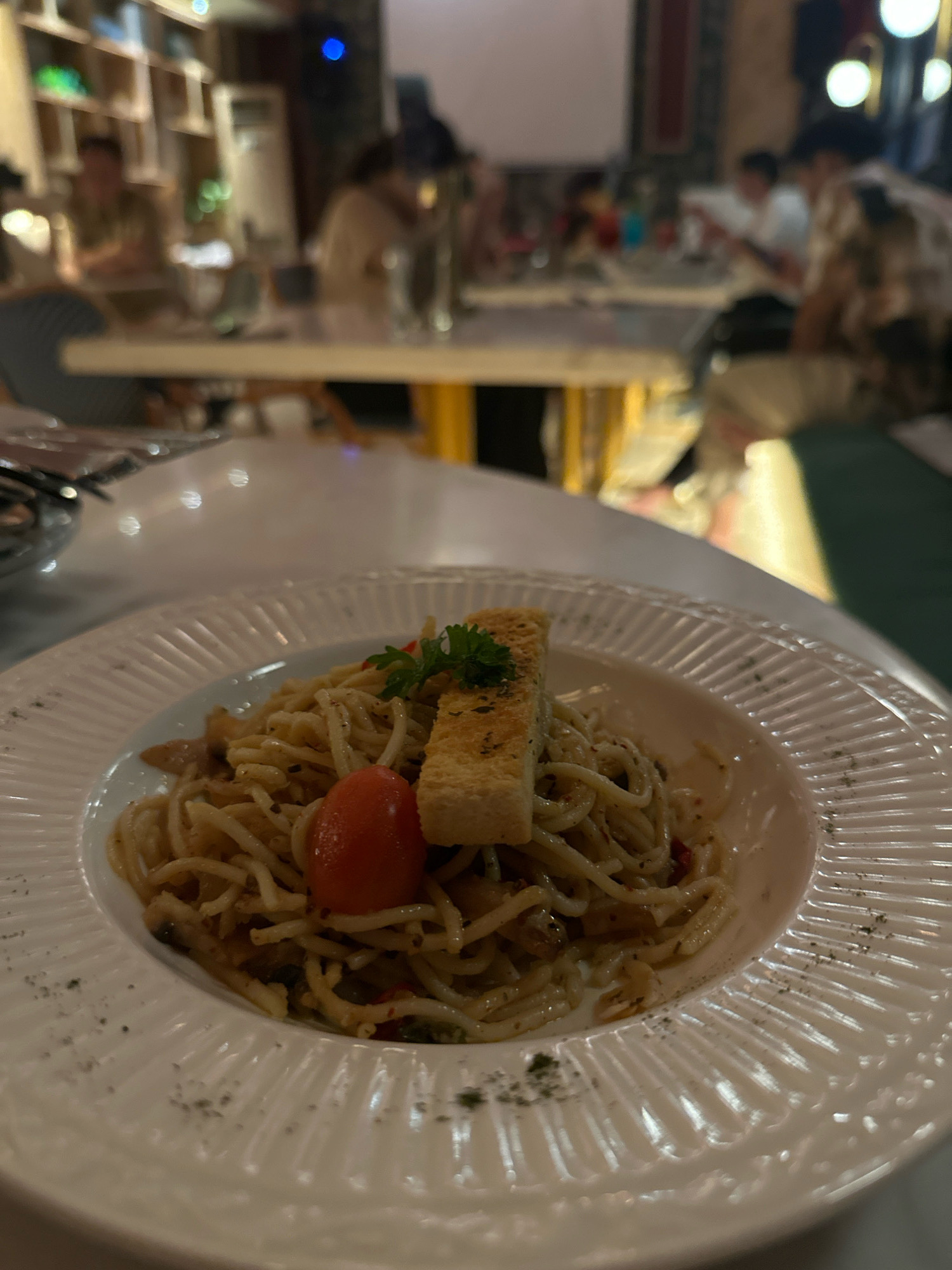 In the foreground, a white, fluted plate holds a serving of spaghetti. The pasta is intertwined and coated in a light sauce, garnished with a single red cherry tomato, a sprig of parsley, and a piece of toasted bread. The background is a blurred depiction of a restaurant interior, suggesting a dimly lit atmosphere with people seated at tables. The overall impression is a close-up shot of a meal in a casual dining setting. This scene is located in the city of Medan, Indonesia, as indicated by the context of the image.