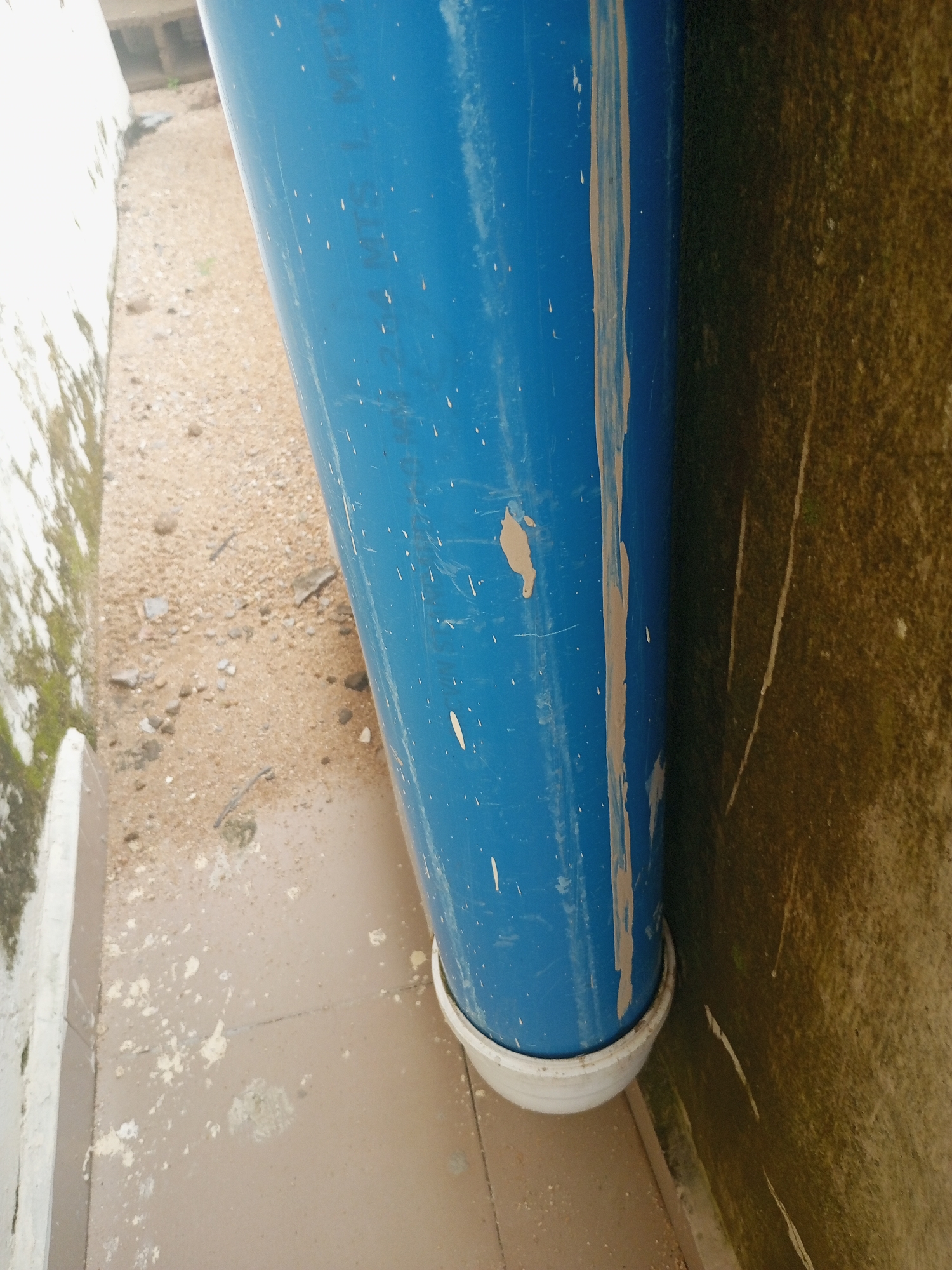 A tall, blue cylindrical object, possibly a pipe or a tank, stands vertically in an outdoor area. The object is speckled with white marks and streaks of a tan, muddy substance. It rests on a white circular base at the bottom, which is partially submerged in a puddle of muddy water. The ground around the base is a mixture of sandy soil, small stones, and debris, with some white splatters, likely paint or dried mud, on the light brown paved surface. To the left of the blue object is a low, white wall with some green mossy growth on its surface. To the right, a textured, dark olive-green wall with faint white streaks runs vertically. The environment appears to be an outdoor urban setting in Jalingo, Nigeria, characterized by the presence of concrete structures and a generally unkempt ground. There are no people or animals visible in the image.