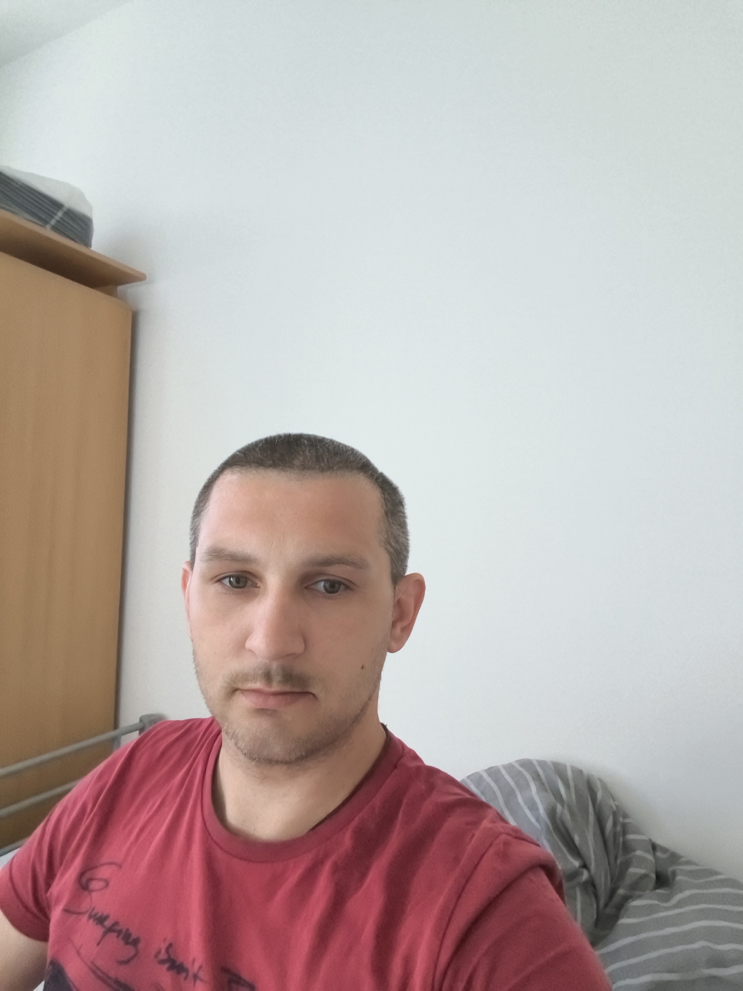 The image is a selfie taken indoors, likely in a bedroom. A man with short brown hair and stubble is looking at the camera. He is wearing a red t-shirt with some text and graphics on it. To his left is a wooden wardrobe or cabinet, and on his right, part of a bed with grey and white striped bedding is visible. The wall behind him is plain white. There are no specific identifying features of Wolgast, Germany visible in the image.