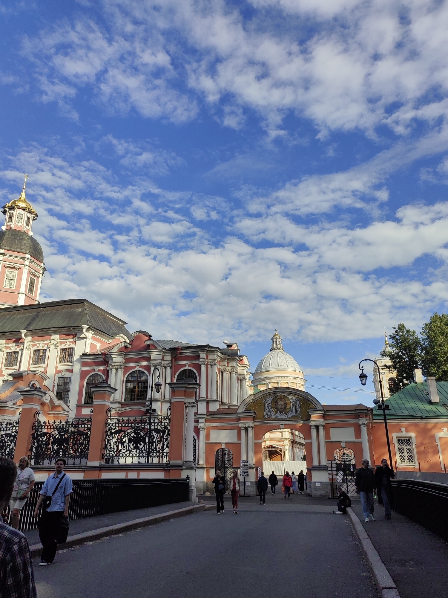 The image depicts an exterior view of a grand, pink and white building with golden domes, likely a religious or historical site, set against a bright blue sky with scattered white clouds. The architecture suggests a Russian Orthodox church or monastery, possibly in Saint Petersburg given the context. The sun is shining, indicating it's daytime, likely late afternoon or early evening due to the shadows.
Several people are visible, walking on a paved walkway in front of the building. They are dressed casually, suggesting they are tourists or visitors. One person in the foreground wears a blue shirt and dark pants, carrying a bag. Others are further down the path, appearing to move towards or away from the entrance. The overall atmosphere seems peaceful and ordinary, with no indication of a specific event.
The building features ornate architectural details, including arched windows, decorative columns, and intricate metal fencing. A prominent archway, adorned with a mosaic image, serves as an entrance. A lamppost stands near the archway, adding to the historical ambiance. The surrounding area appears to be well-maintained. There is no visible text in the image.