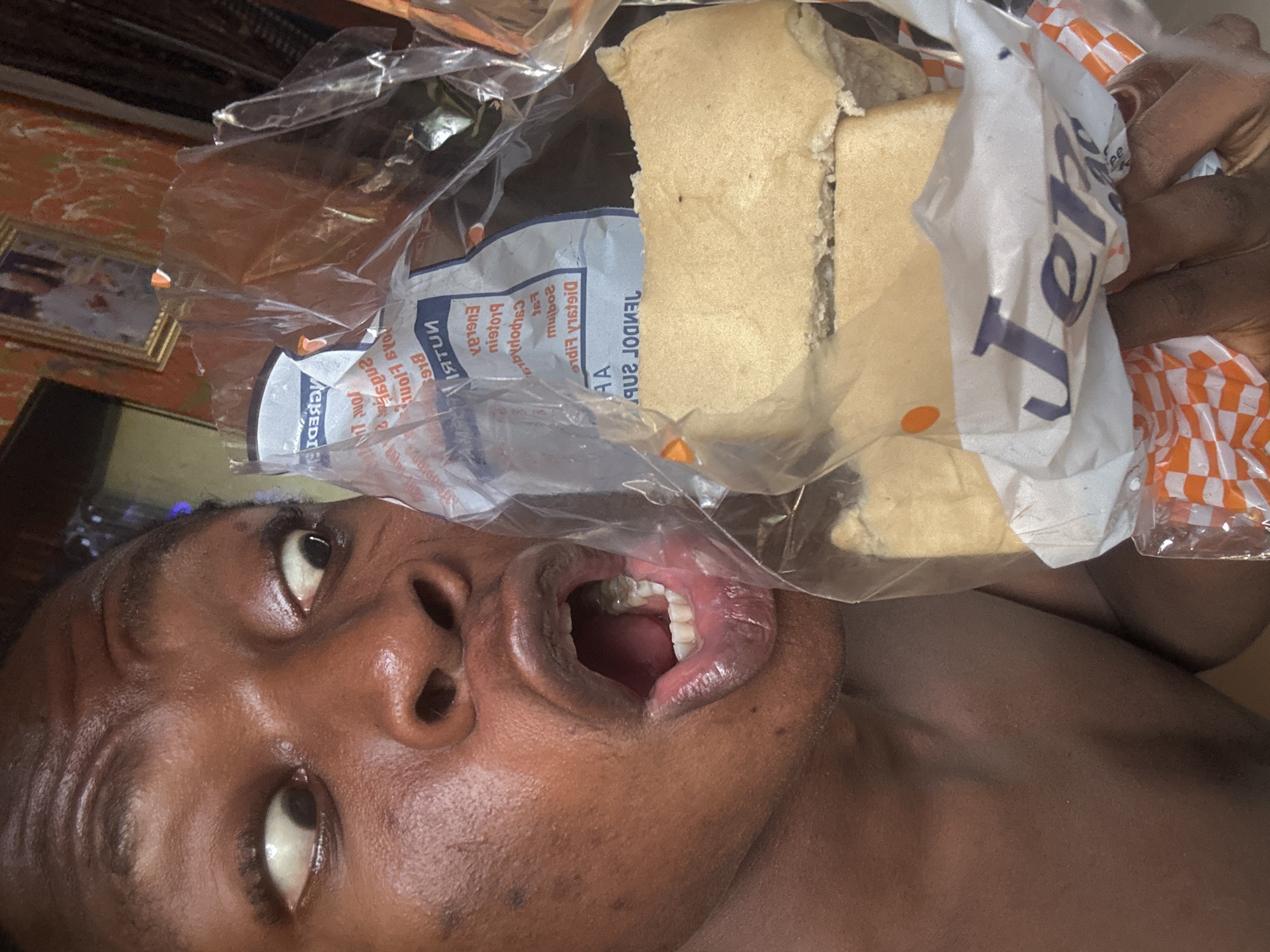 A close-up, tilted selfie features a dark-skinned person with eyes looking upward and mouth open as if about to eat something. The person's face exhibits fine lines and open pores. In front of the person's mouth, a clear plastic bag contains two rectangular slices of light tan-colored food, possibly bread or a similar item. The bag also displays printed text, including the word "JERK" in large letters. The background includes a glimpse of an interior setting with textured walls and a framed picture. Part of the person's torso is visible, indicating that the person is shirtless.