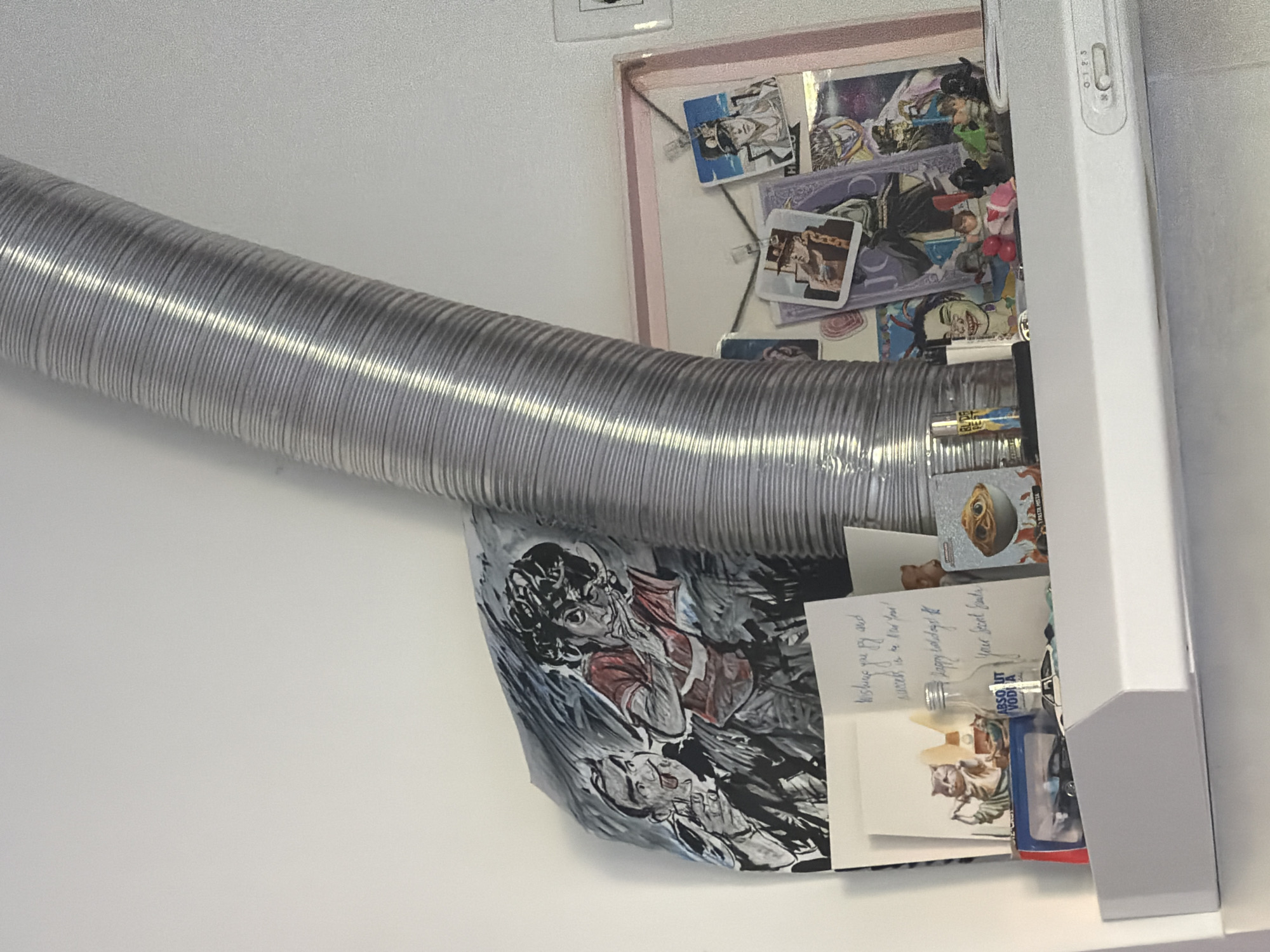 The image shows a close-up view of a kitchen area. A corrugated silver pipe is positioned horizontally across the frame. A white cabinet with a light switch on the right side is filled with various items. These items include what appear to be collectible cards attached to a pink-edged frame, a black and white illustration, greeting cards, and small liquor bottles. The setting appears to be indoors, possibly in a residential kitchen, and it is unlikely that the photograph's location within Podgorica, Montenegro can be determined from the provided imagery.