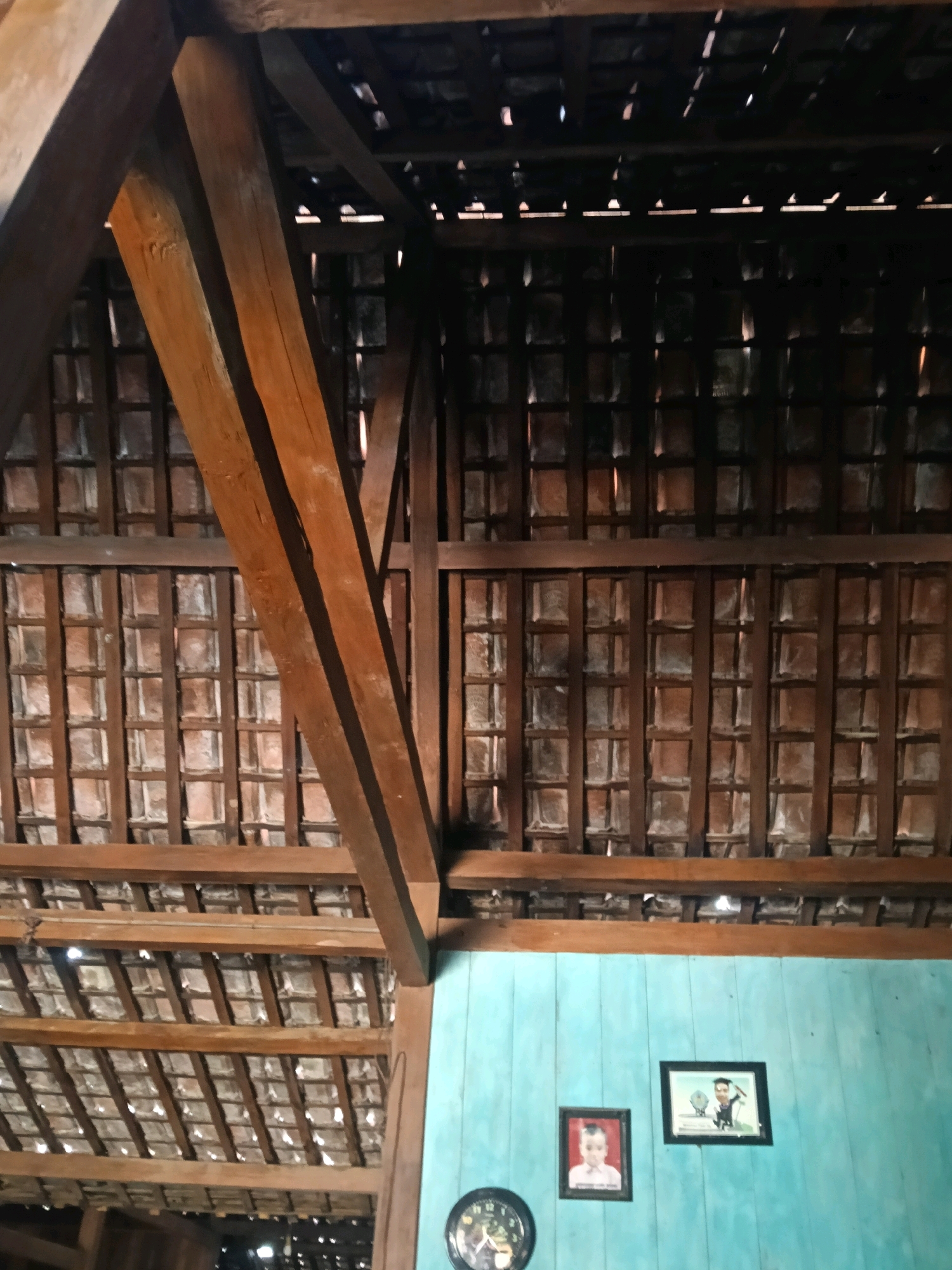 The image shows the interior of a building with a prominent wooden roof structure. The roof is made of clay tiles and supported by large wooden beams. Below the roof, there is a light blue wall. On the wall, there are two framed pictures: one of a child and another of a person in a graduation cap. To the left, a portion of a clock face is visible. The overall impression is of a rustic and simple interior. The location is identified as Nglungger, Indonesia.