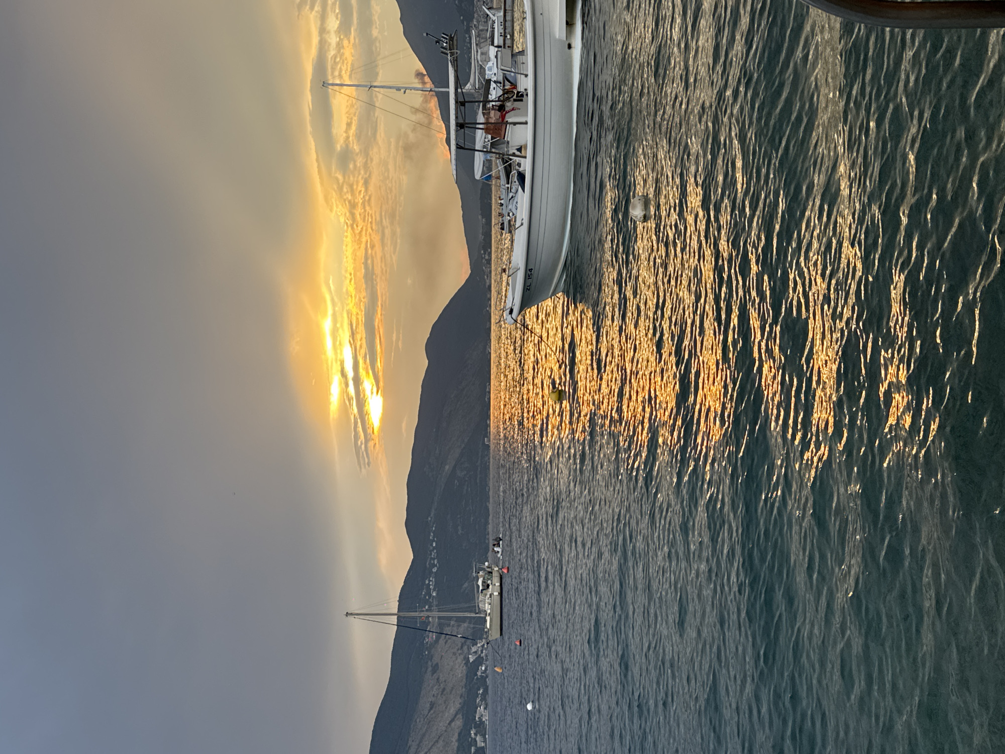 A serene bay scene unfolds at dusk. The sky is a dramatic canvas of grays and oranges, with the sun setting behind a range of dark, silhouetted mountains. The water reflects the warm hues of the sunset, creating shimmering streaks of light. Two boats are visible: a sailboat with its mast reaching towards the sky in the mid-left, and a white motorboat with a canopy in the mid-right. A few buoys dot the water, and a person appears to be seated on the motorboat. The distant shoreline is dotted with lights, suggesting a town or village nestled against the mountains. The overall impression is one of tranquil beauty and the peaceful end of a day.
