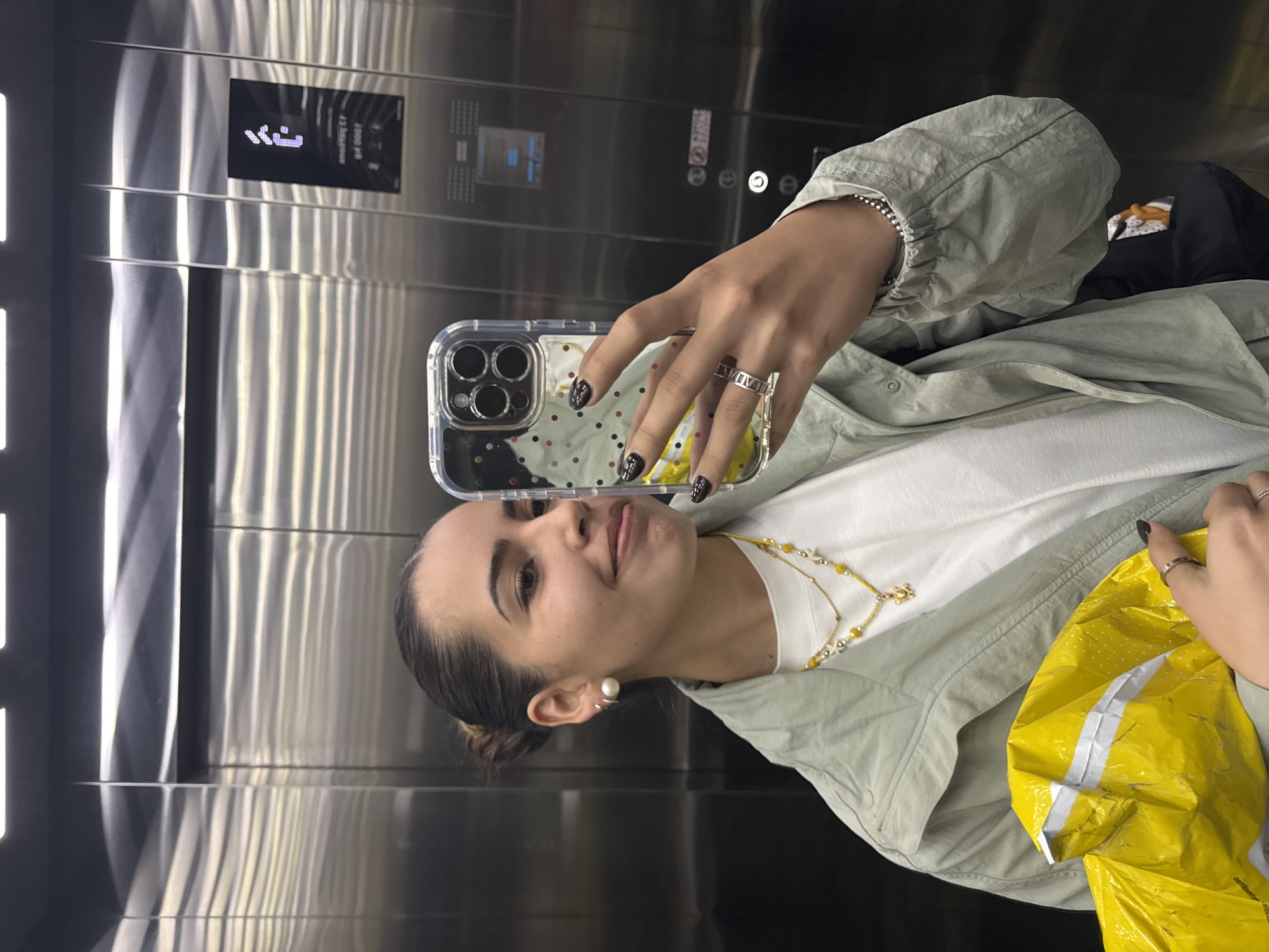 A person is taking a selfie in an elevator. The elevator's interior is stainless steel, with control panels and floor indicators visible. The person in the photo is wearing a white shirt, a light green jacket, and a yellow and white necklace. They have dark hair pulled back, and are wearing rings and earrings. They are holding a smartphone in their hand, which has a clear case. The city of San Bernardino Tlaxcalancingo, Mexico, cannot be confirmed.