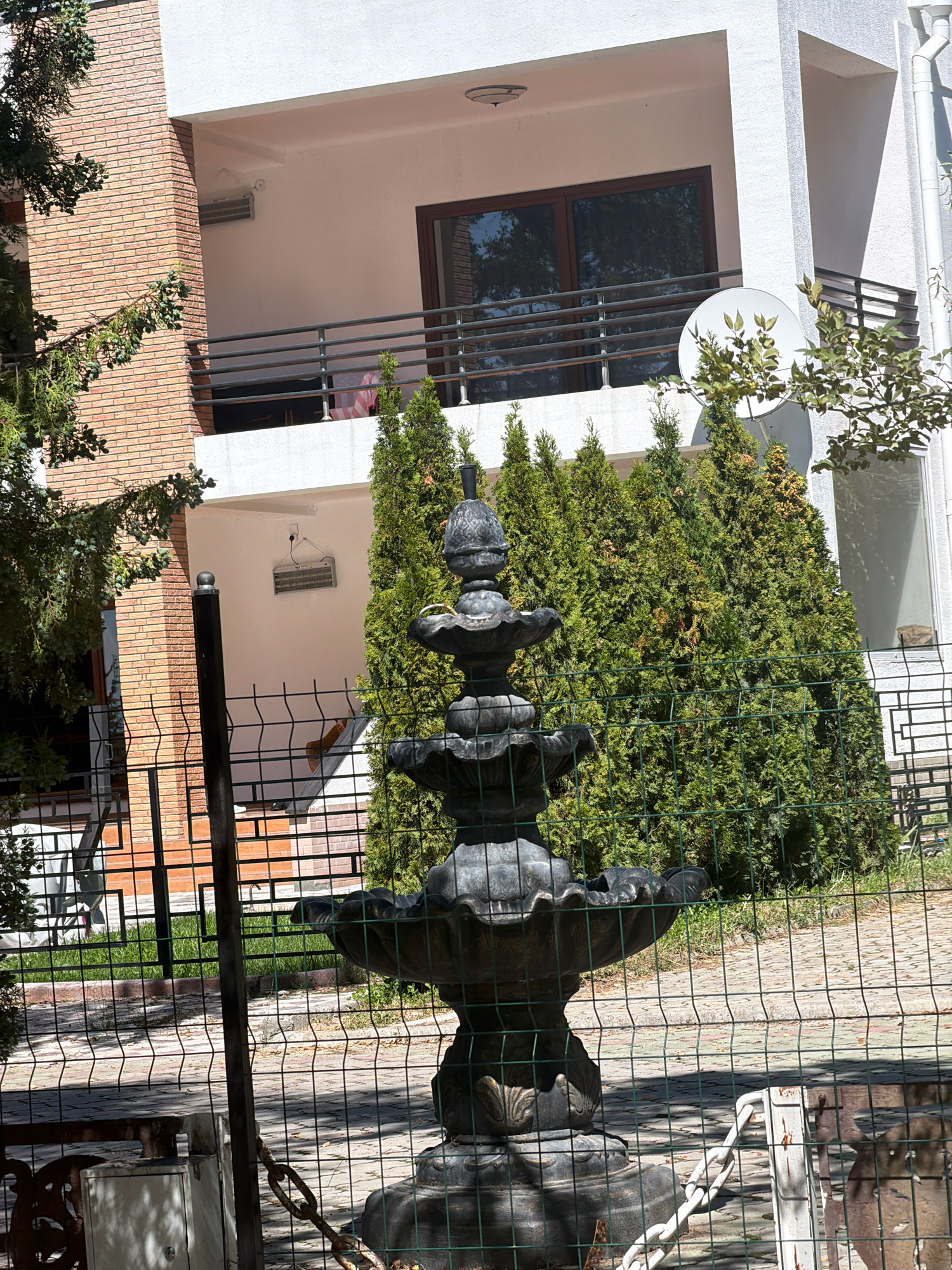 The image depicts a multi-story building with a brick facade on the left and white stucco on the right, suggesting a residential or commercial property in a city. A prominent feature in the foreground is a tiered, dark-colored fountain made of what appears to be concrete or stone, partially obscured by a green wire fence. Several tall, conical evergreen trees are planted behind the fountain, forming a dense green backdrop. A balcony with a railing is visible on the second floor of the building. The ground in front of the building is paved with light-colored stones, and there is a patch of green grass to the left of the fountain. The overall scene suggests an outdoor area, possibly a garden or courtyard, attached to the building. The specific location in Бактуу-Долоноту, Kyrgyzstan, is inferred but not directly visually confirmed within the image itself.