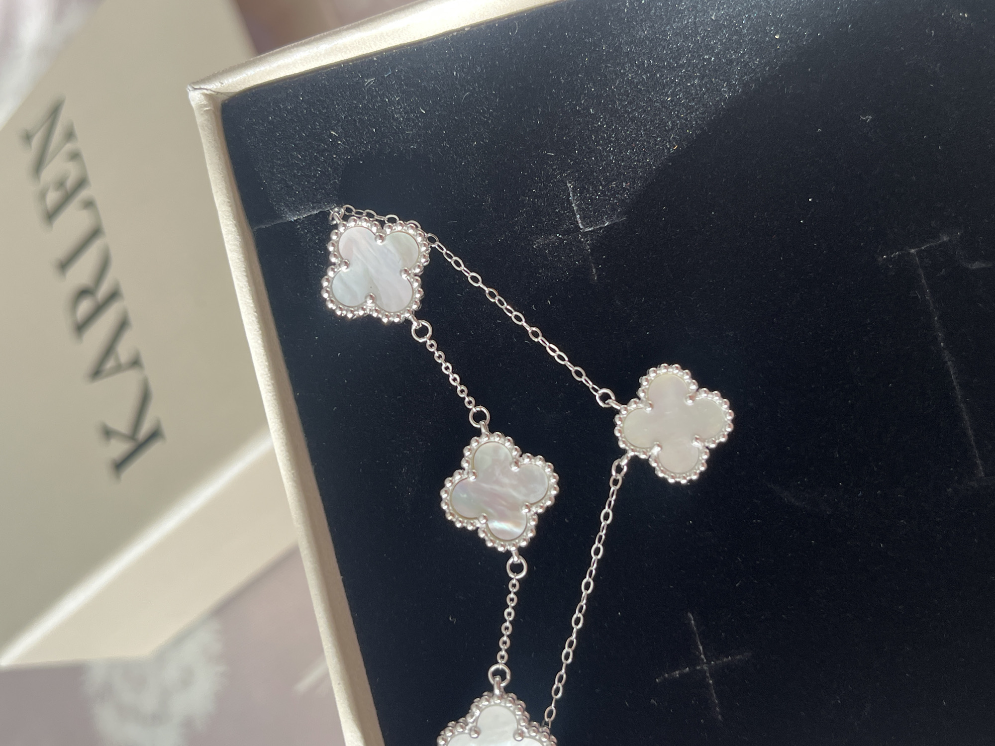 The image shows a silver necklace with white clover-shaped pendants displayed inside a dark box. A box with the word "KARLEN" printed on it is partially visible to the left.