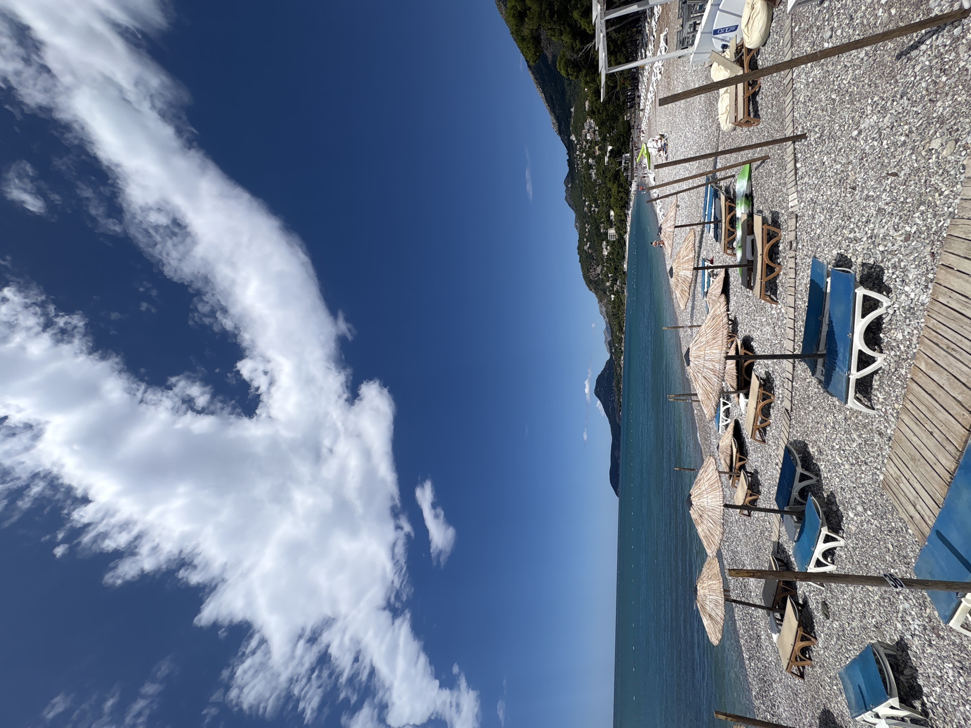 A wide-angle, full shot captures a pebble beach in Bar, Montenegro, under a bright blue sky with scattered white clouds. The foreground features rows of empty lounge chairs and straw umbrellas, some with wooden frames. Wooden walkways are laid out on the pebble surface. To the left, the turquoise sea stretches towards the horizon, with a distant, dark, mountainous island. Along the coastline, the land rises into green, forested hills dotted with buildings. Further down the beach, a few people are visible, some sitting on chairs or walking. The overall scene depicts a serene, undeveloped beach preparing for visitors.