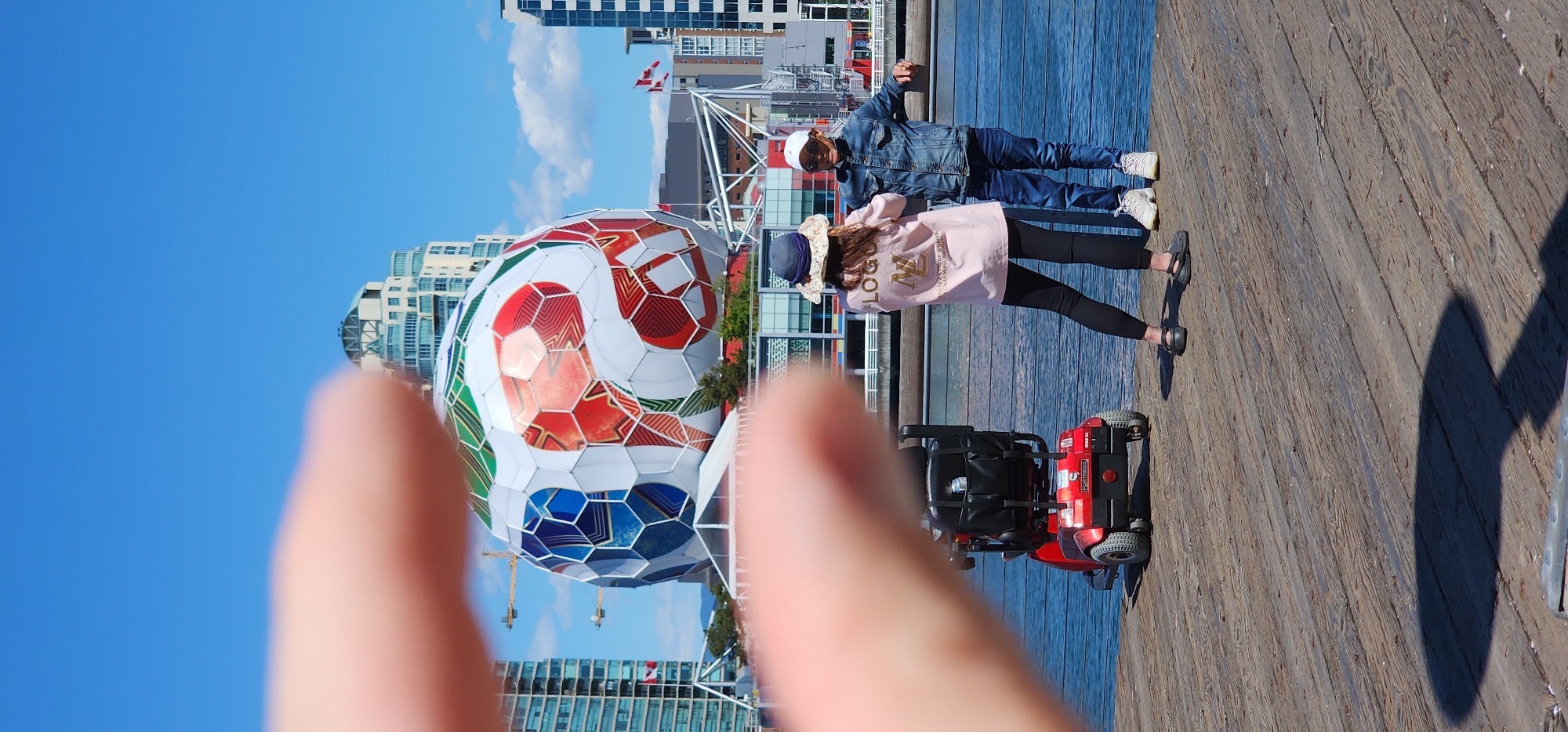 In an outdoor urban scene on a sunny day, two people stand on a wooden boardwalk near a body of water. A red mobility scooter is parked to their left. In the background, a large, geodesic sphere decorated with a soccer ball pattern is visible, along with several modern buildings and construction cranes. The sky is clear blue with some scattered clouds. One person, facing away from the camera, wears a light pink long-sleeved shirt with gold lettering, black leggings, and a patterned hat. The other person, also facing away, wears a blue denim jacket, blue jeans, a white baseball cap, and white sneakers. Their hands are slightly extended as if they are talking or gesturing. The scene is framed by a blurred foreground of a human finger, suggesting a first-person perspective.