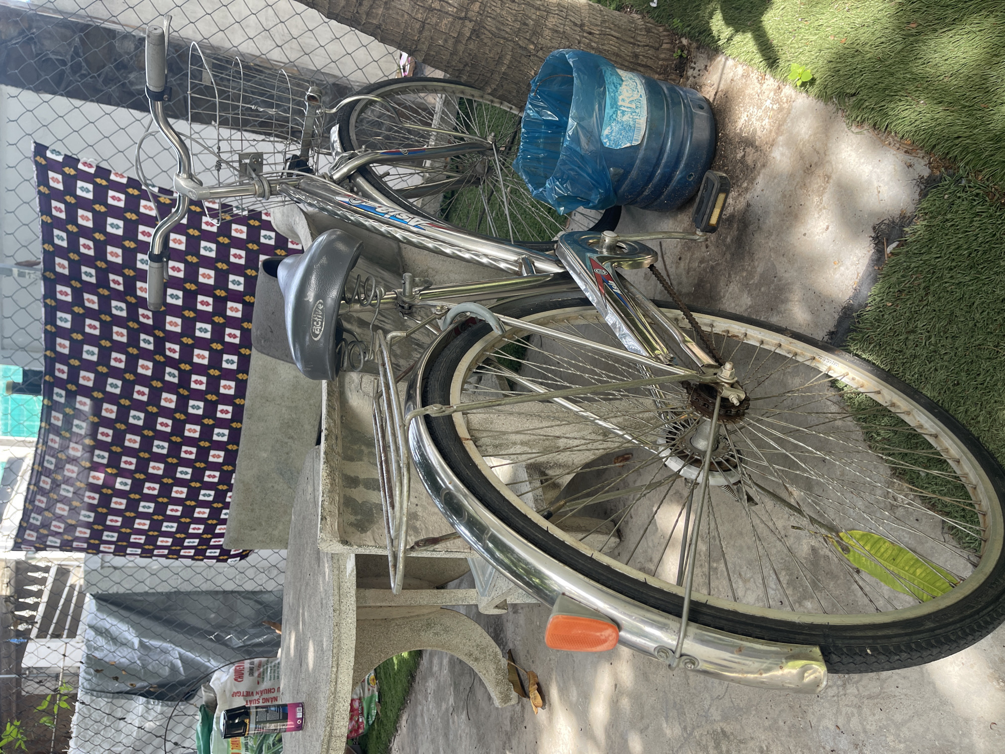 The image shows a silver bicycle with a black seat and a wire basket, leaning against a chain-link fence. A blue plastic trash can is next to the rear wheel of the bicycle. A patterned cloth hangs on the fence behind the bicycle. In the foreground, a concrete table and benches are visible. The ground is a mix of concrete and grass. A small green plant is growing on the grass. The scene appears to be outdoors in a residential or public area. The location is described as Phu Thuy Ward, Vietnam.