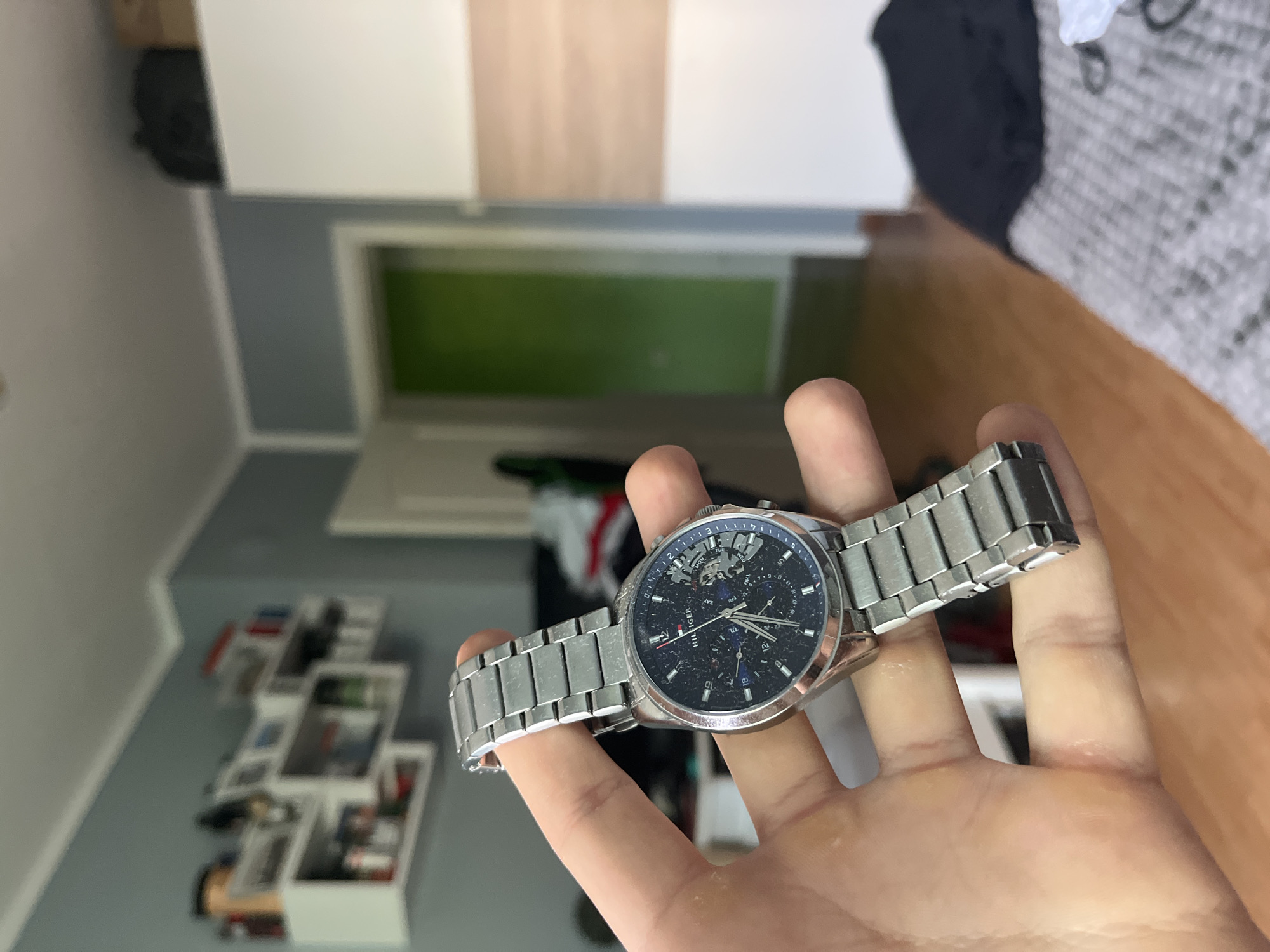 A close-up, rotated shot depicts a person's hand holding a silver-colored wristwatch. The hand is positioned centrally, presenting the watch. The wristwatch features a metal link band and a dark blue or black dial. Visible text on the dial identifies the brand as "HILFIGER." The watch face is multi-functional, displaying a day-of-the-week sub-dial (with "SUN, MON, TUE" visible), a date sub-dial (showing "18"), and another sub-dial that appears to be a 24-hour or moon phase indicator. Parts of the watch's internal mechanisms, including gears, are visible through a semi-skeletonized section of the dial, particularly around the upper half. The watch crystal appears somewhat speckled or dusty.
The background shows an indoor setting, likely a room in a residence in Pokrovnik, Bulgaria. To the left, there's a wall painted light grey, with a collection of framed pictures or photos arranged on it. In the mid-ground, a doorway with a bright green interior is visible. Furniture, including what appears to be a light-colored wooden surface (possibly a desk or cabinet) and parts of a bed or sofa with patterned bedding, are also present in the blurred background. The overall lighting suggests it is daytime.